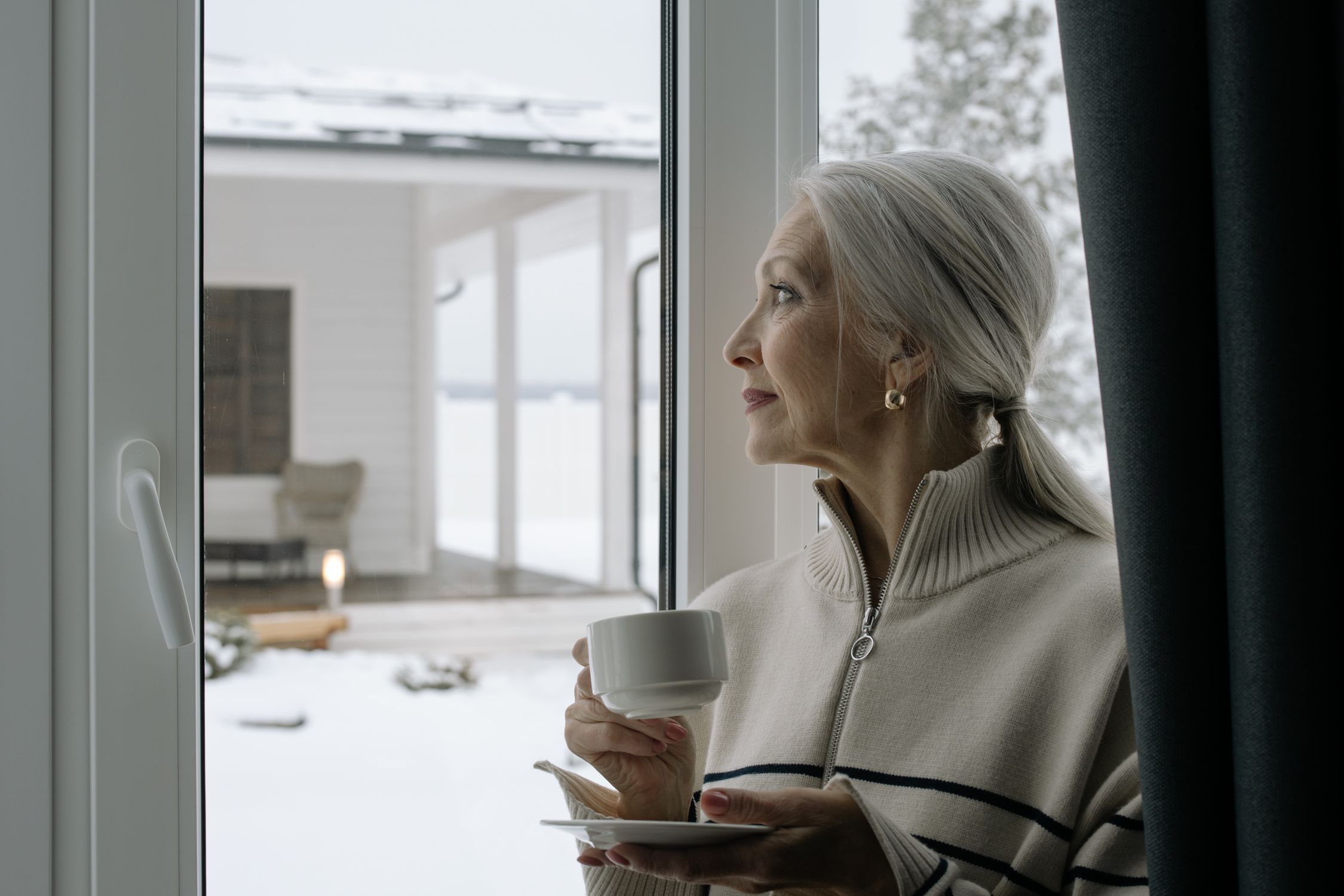 Woman over 60 with grey hair holding coffee cup looking out window at winter scene