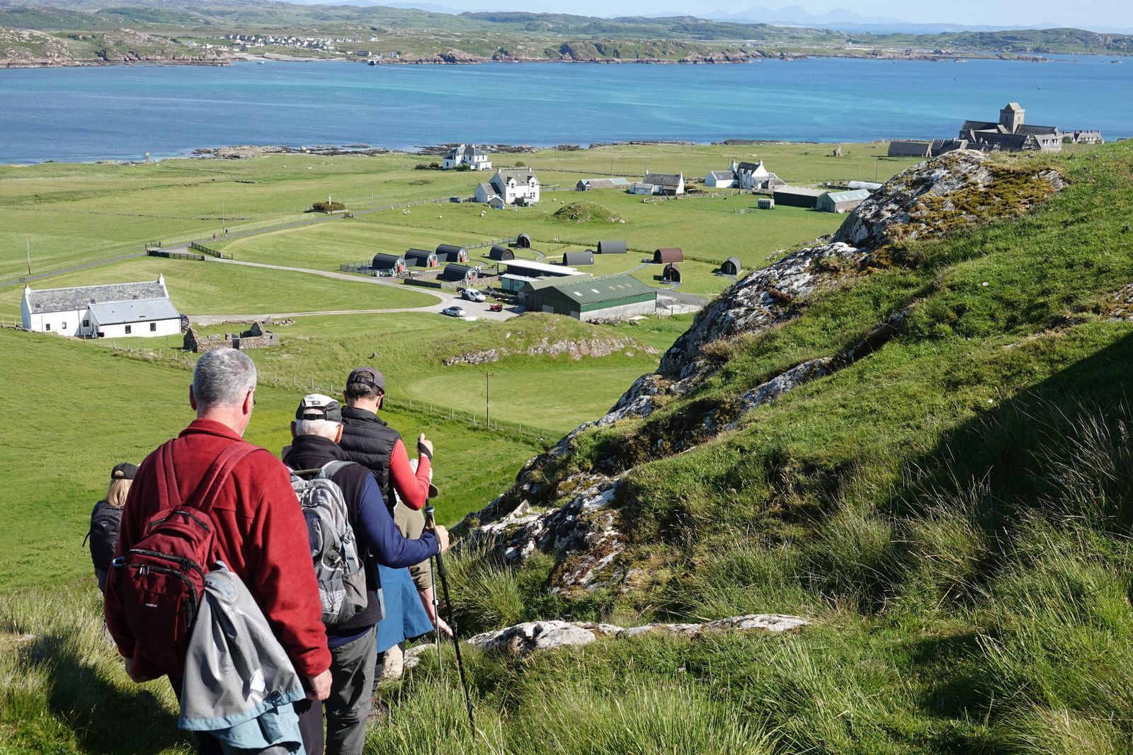 Group of grey-haired hikers descending a green hillside toward a Scottish coastal village on a sunny day