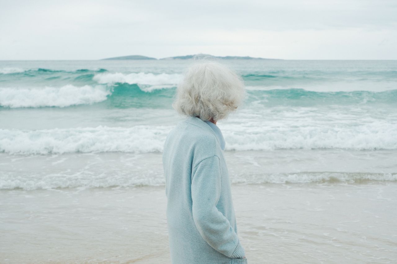 White-haired woman standing alone on an overcast beach, facing the ocean waves