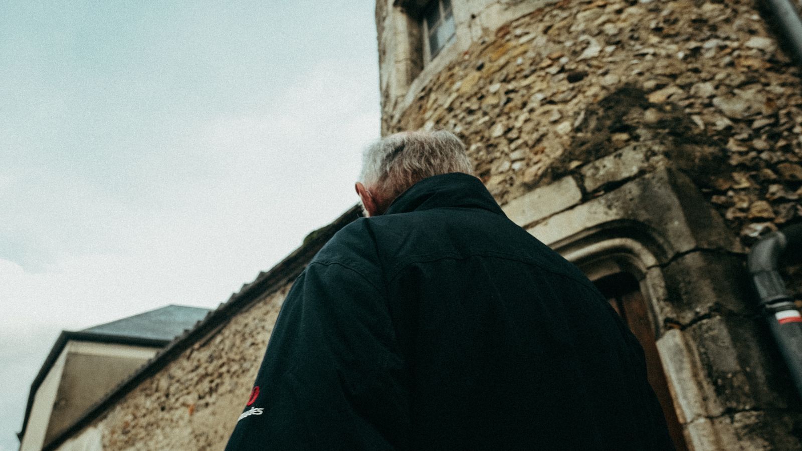 Elderly man in a dark jacket walking past a stone building on a quiet street
