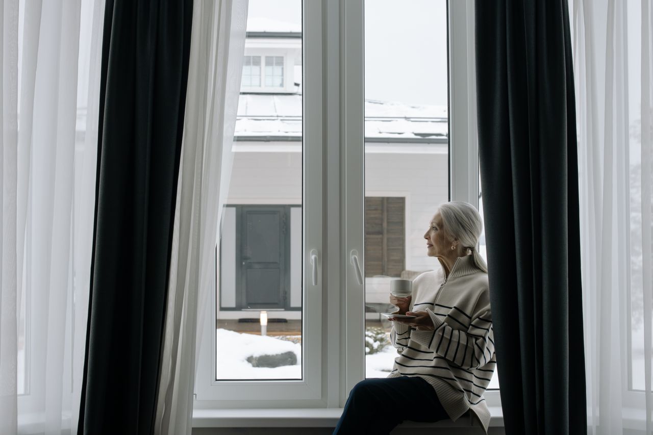 Silver-haired woman holding a cup by a window in winter, looking outward in quiet thought