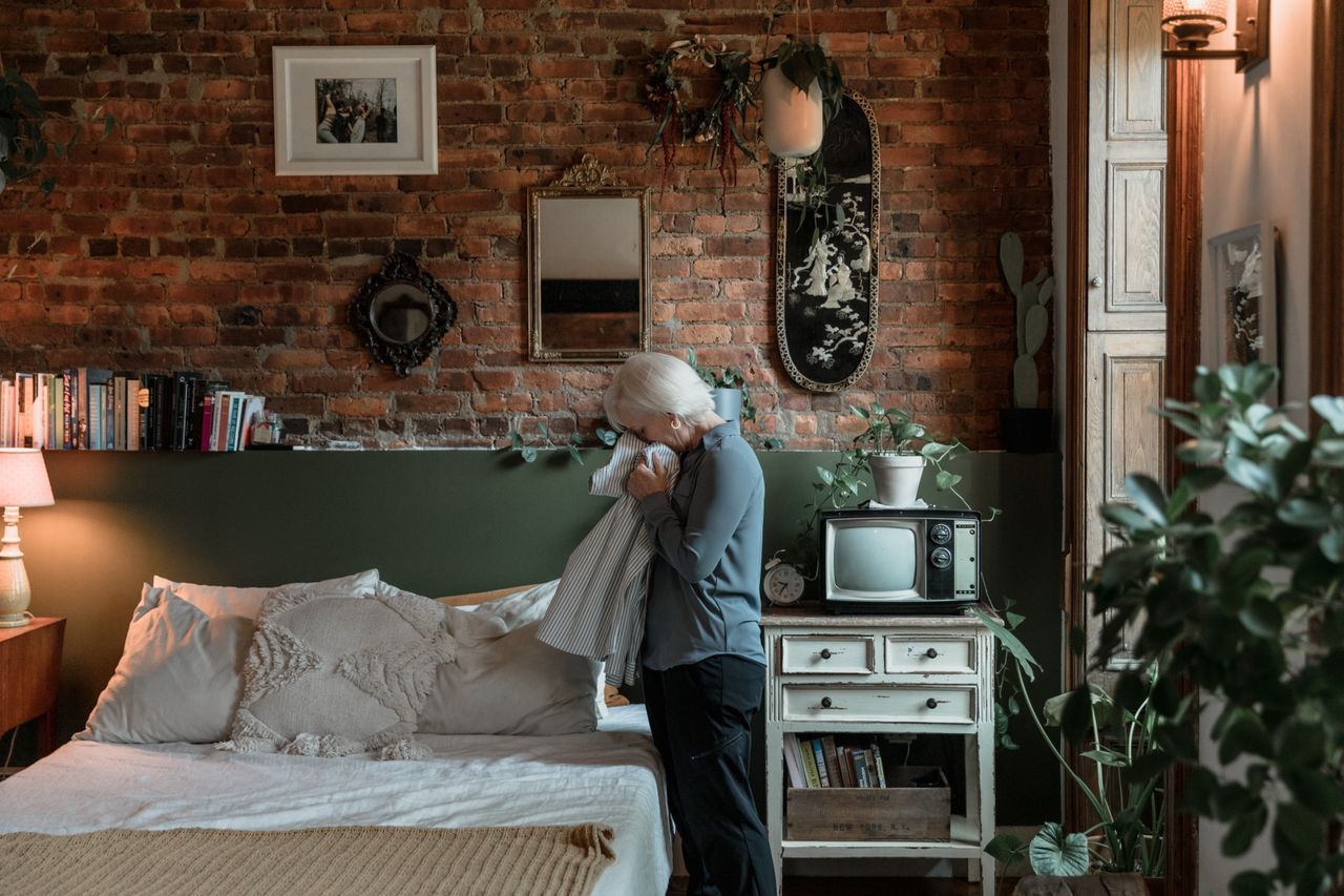 Older woman sitting in thought in a warmly lit bedroom with brick walls