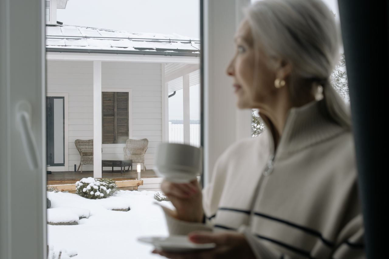 Senior woman holding a ceramic cup and gazing out a window at a snowy porch