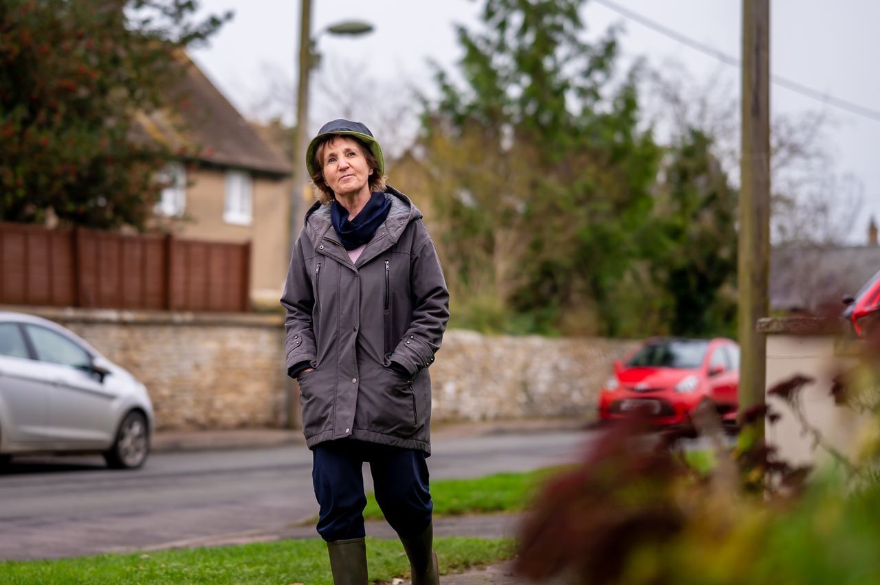 Woman in her sixties standing alone on a quiet residential village street in autumn, looking outward