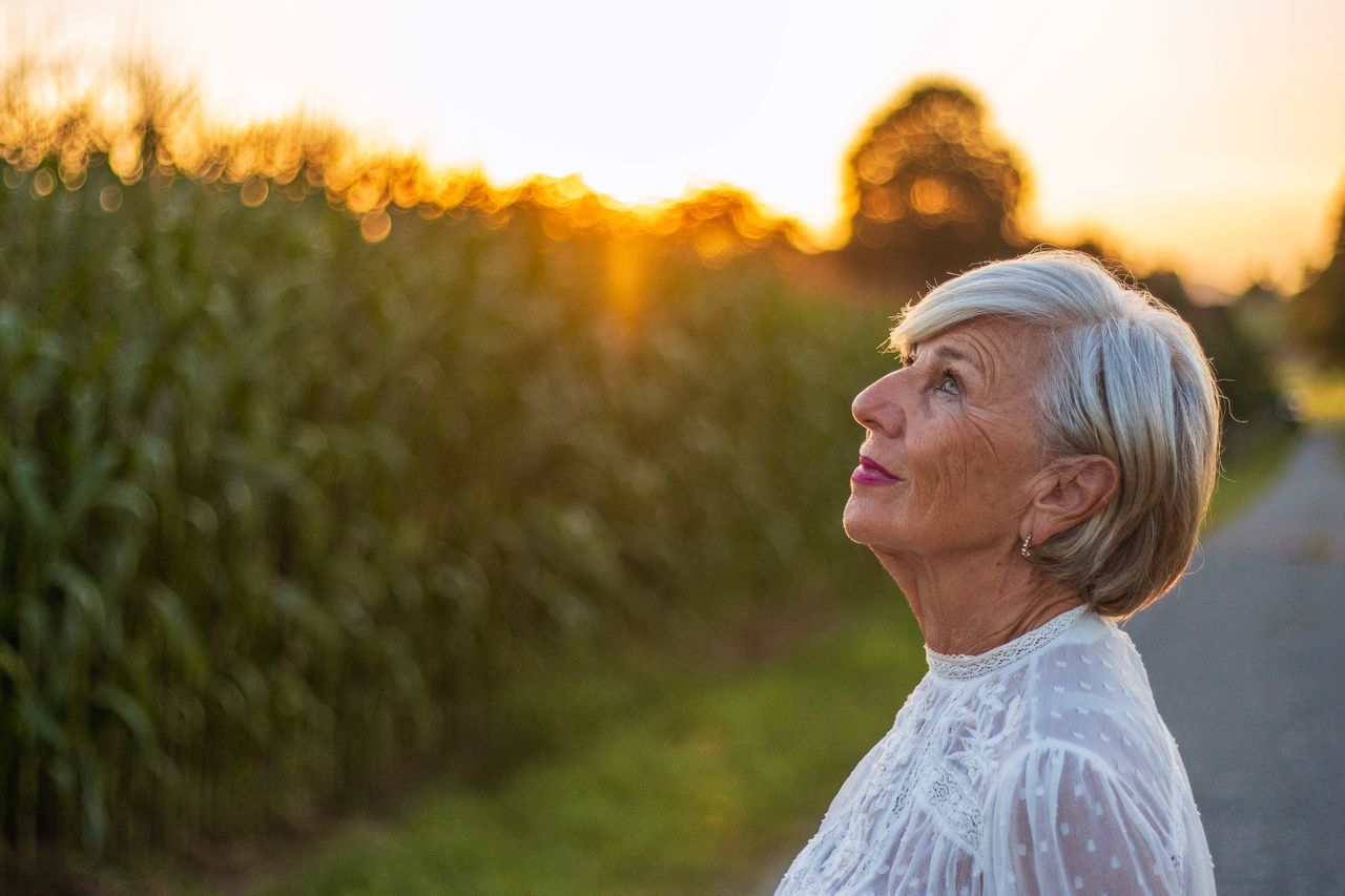 Silver-haired woman in her seventies looking upward beside a green field at golden hour
