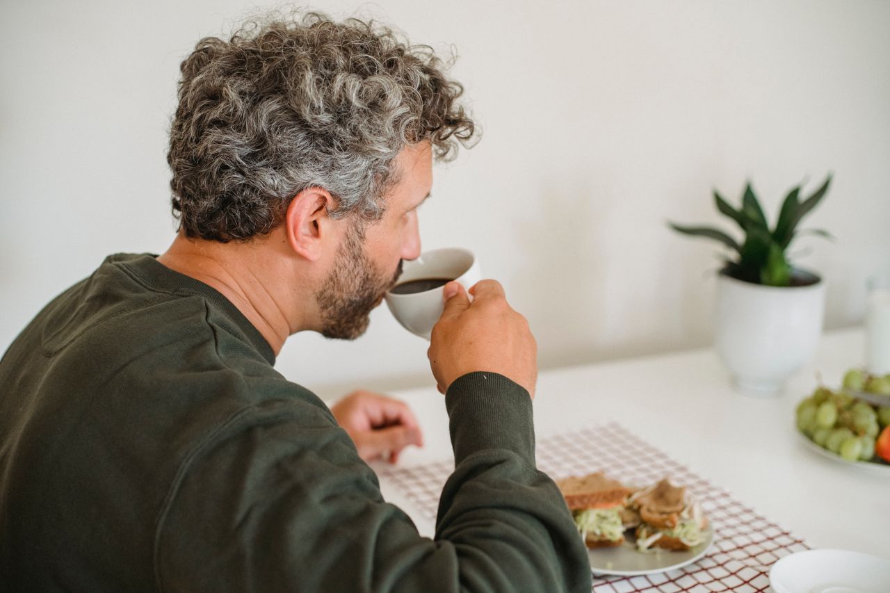 Gray-haired man with a beard sitting at a kitchen table with coffee, looking down thoughtfully