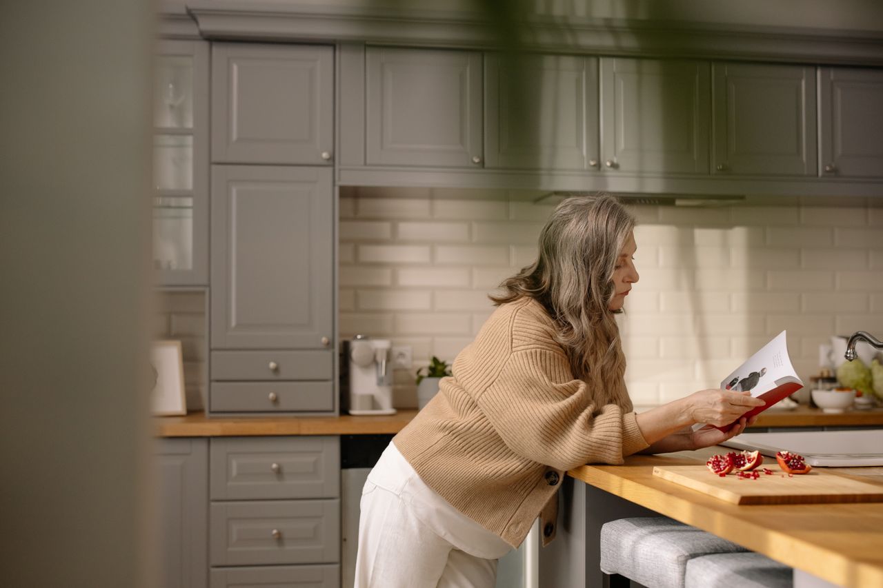 Woman with long gray hair leaning against a kitchen counter reading a book in warm natural light