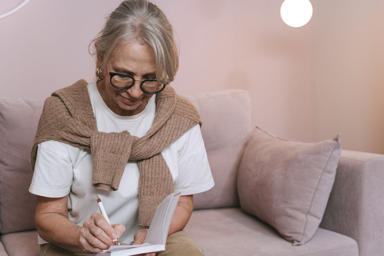 Woman over 60 with glasses writing in a notebook on a sofa at home