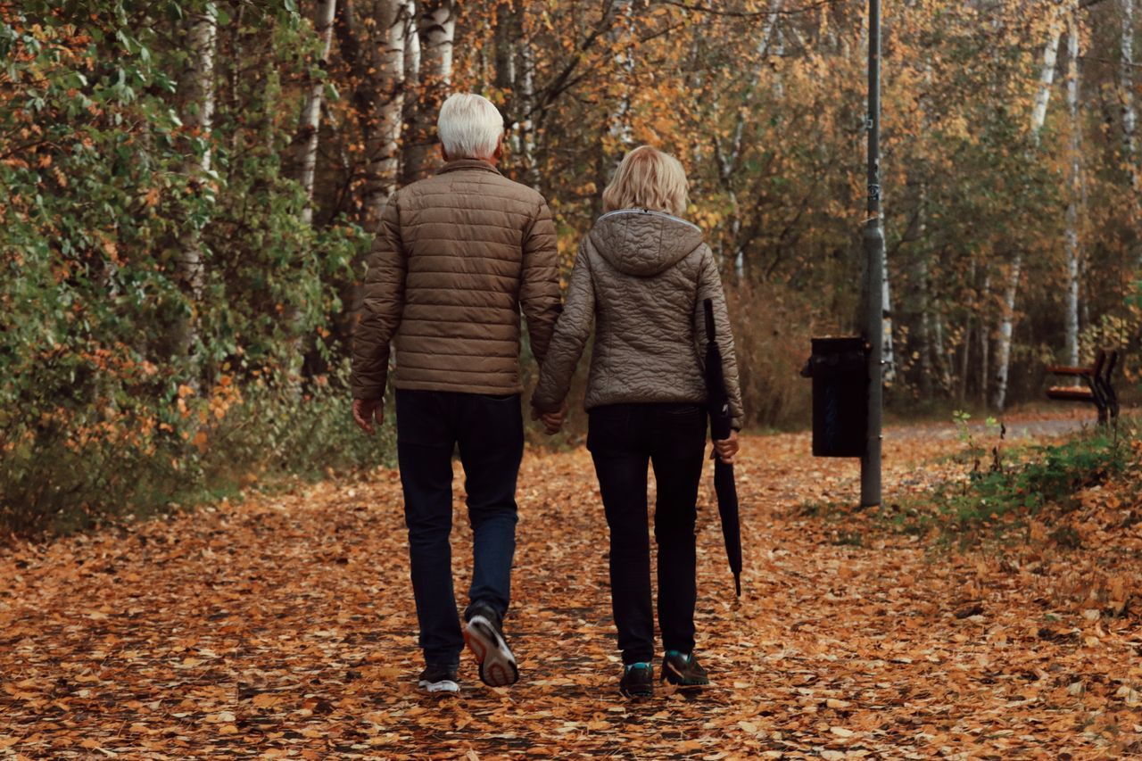 Older couple walking hand in hand along a leaf-covered autumn path through birch trees