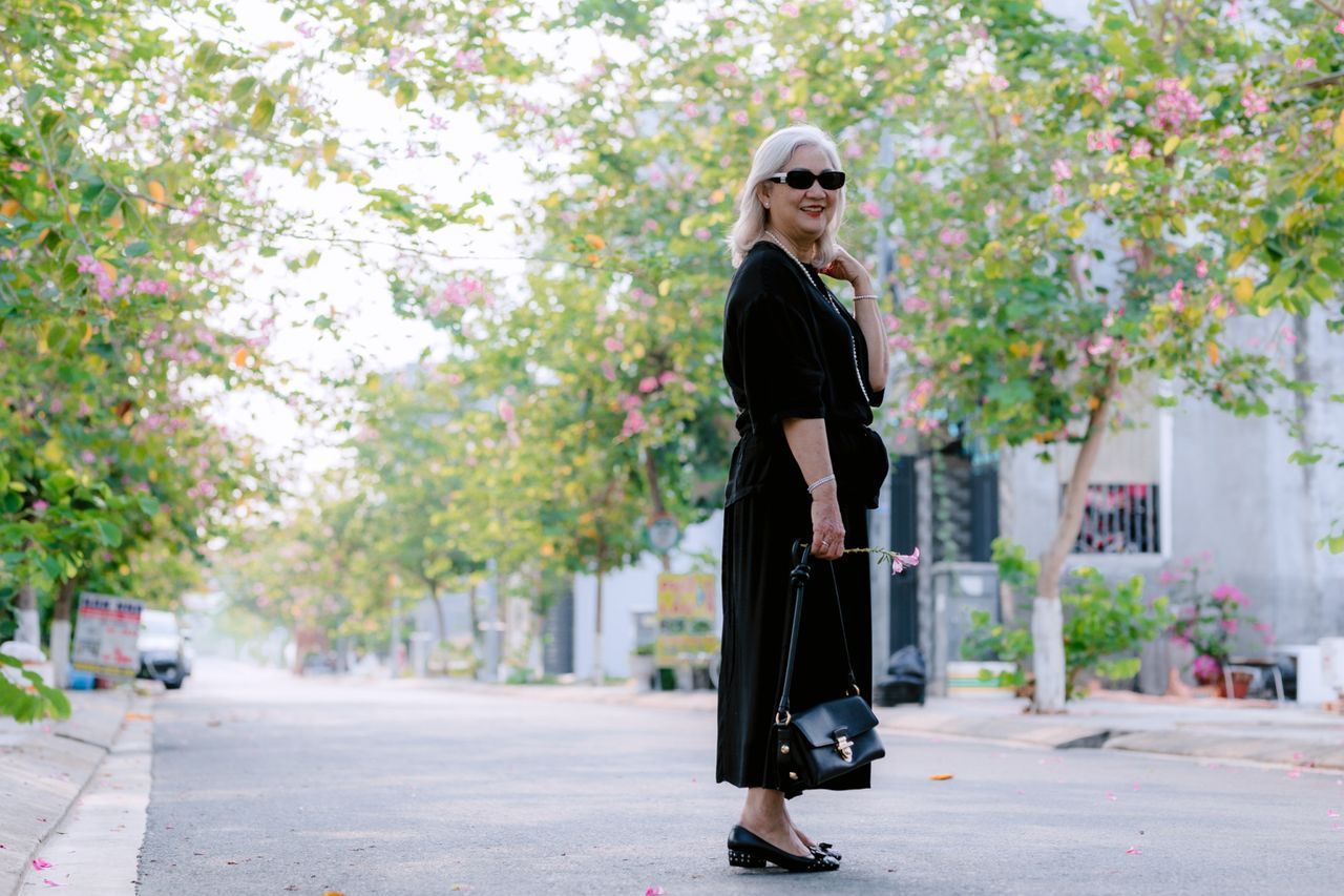 Older woman with white hair in a black dress pausing on a quiet tree-lined street with pink blossoms