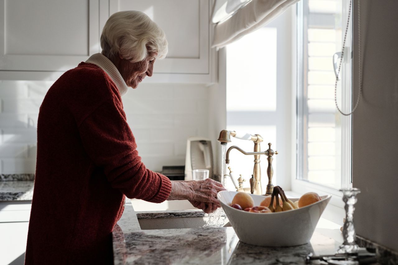 Elderly woman with white hair washing fruit at a sunlit kitchen sink in a burgundy sweater