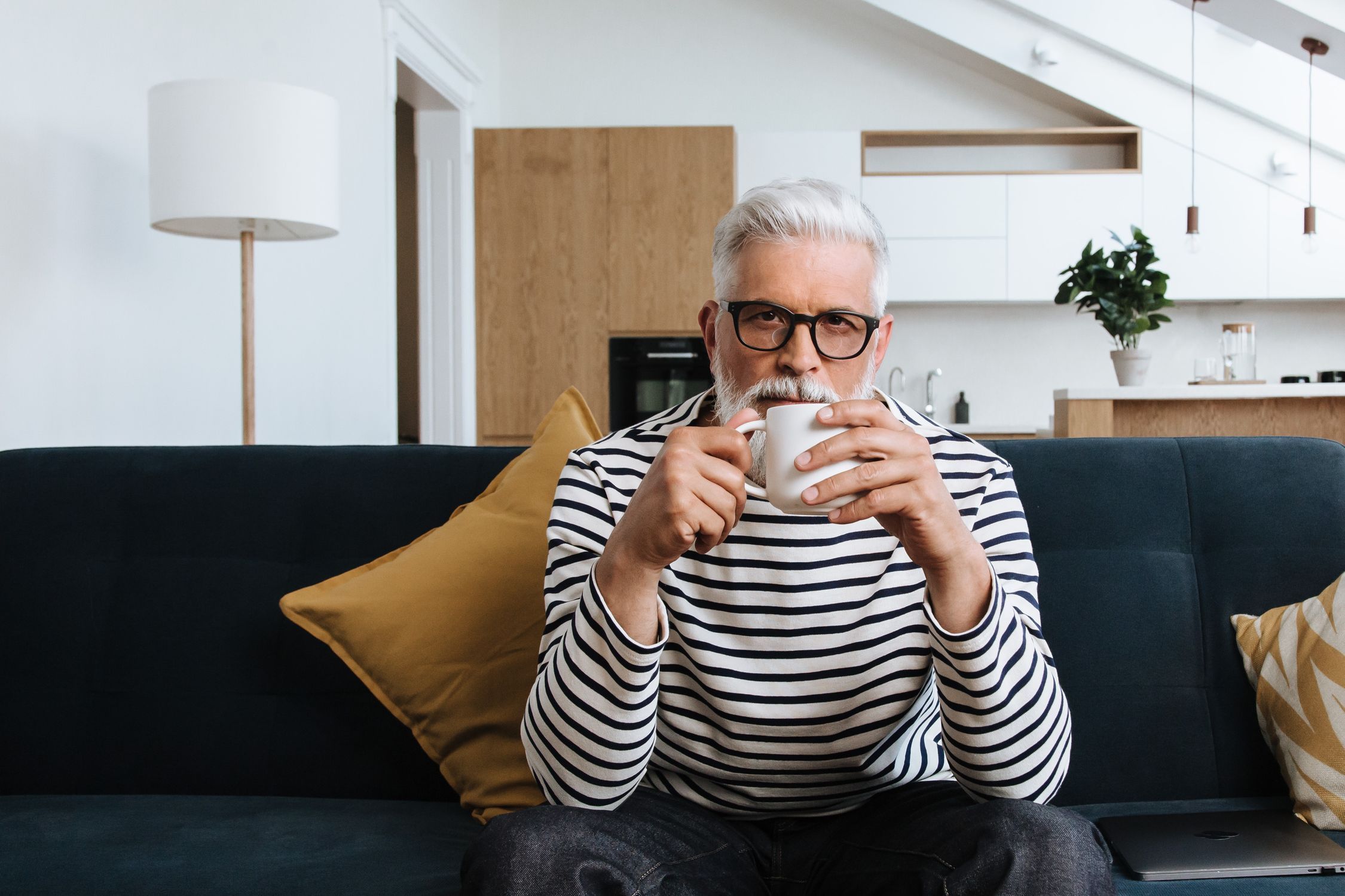 Grey-haired man with glasses sitting on a couch at home, drinking coffee and looking thoughtful