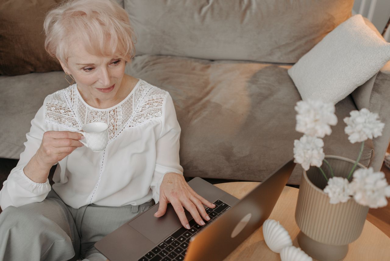 Senior woman with short hair holding a tea cup while browsing a laptop at a home table