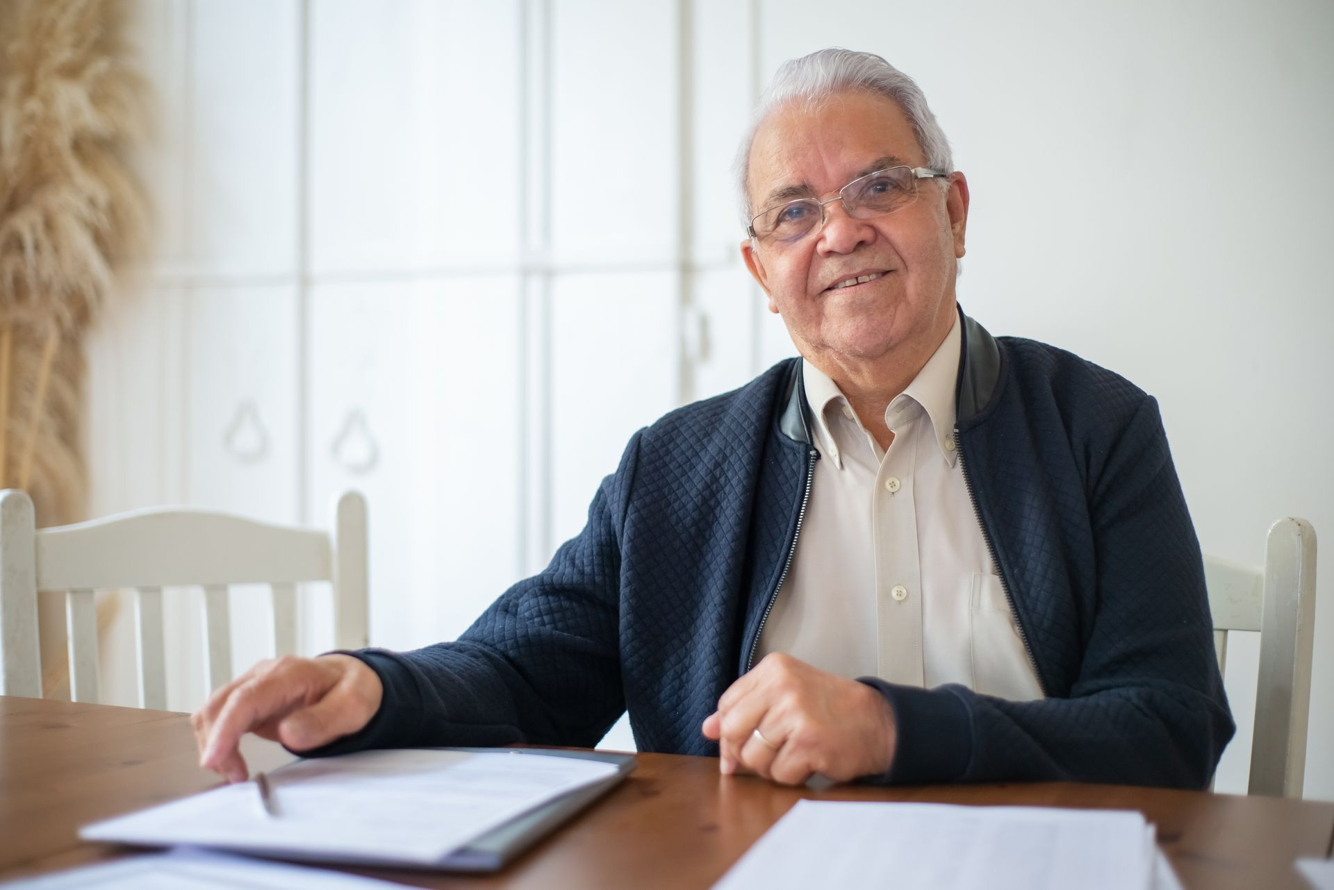 Senior man with white hair and glasses seated at a wooden dining table with paperwork