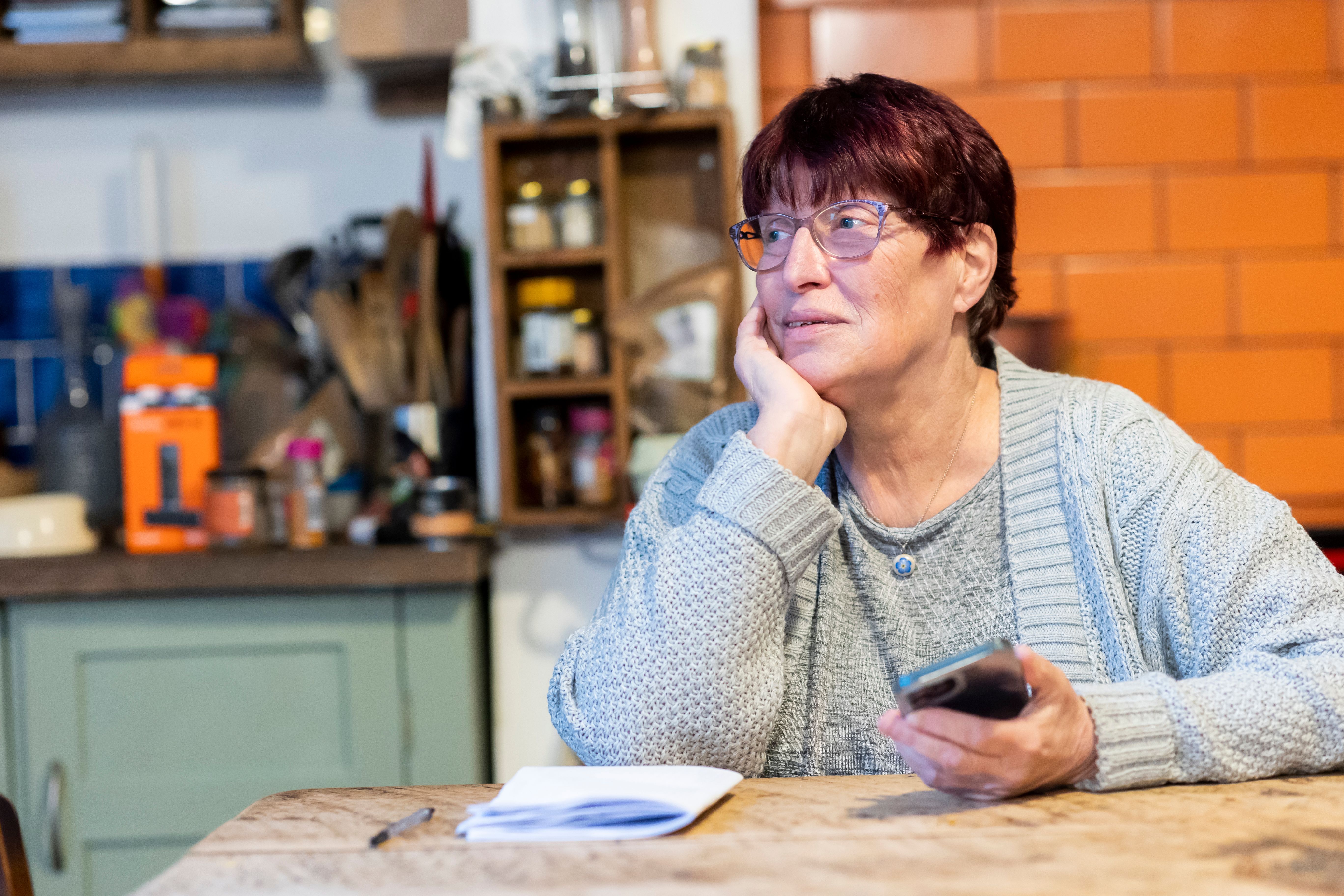 Older woman with glasses holding smartphone at wooden kitchen table, chin resting on hand