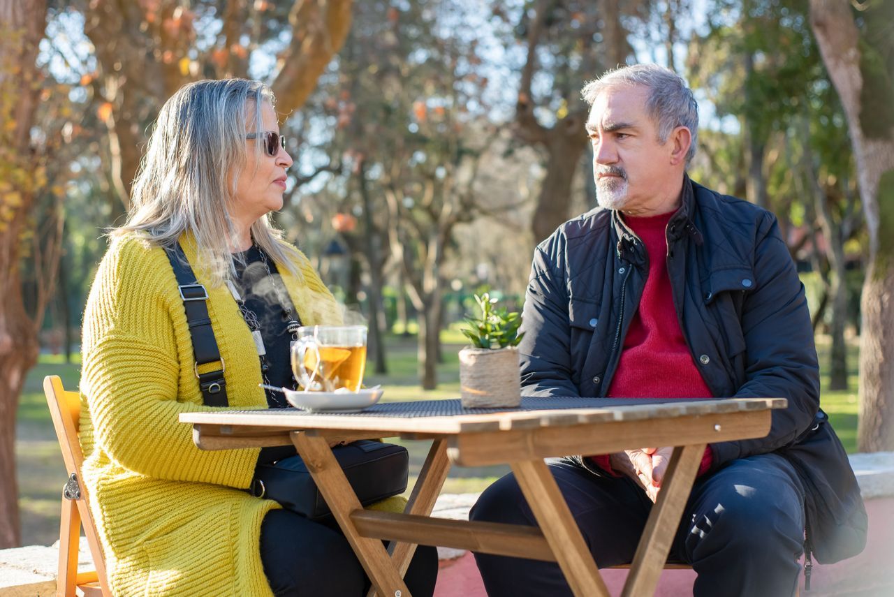 Mature man and woman in conversation at an outdoor table in a sunny park with tea