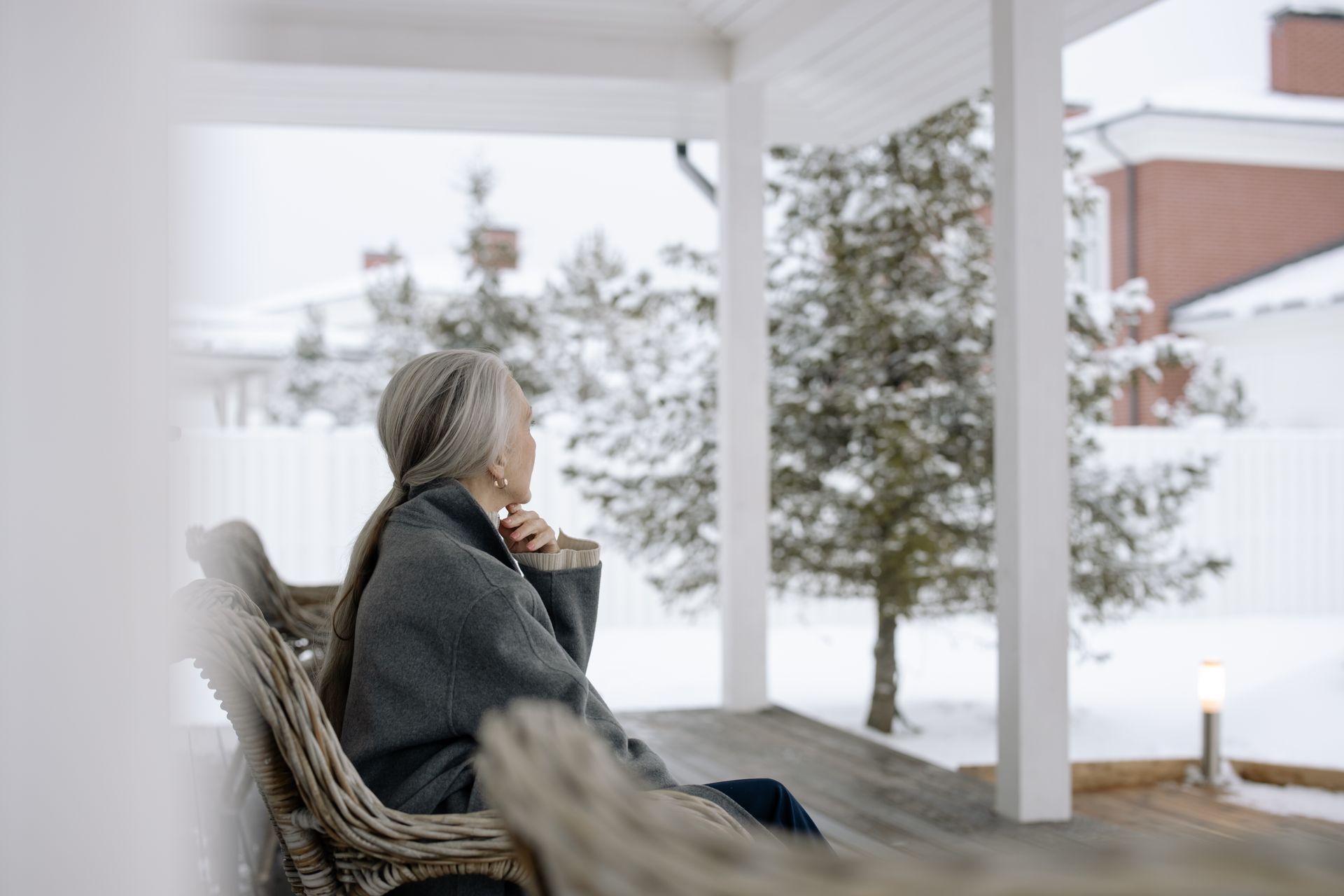 Woman with silver hair sitting on a porch in winter, looking outward at a snowy garden