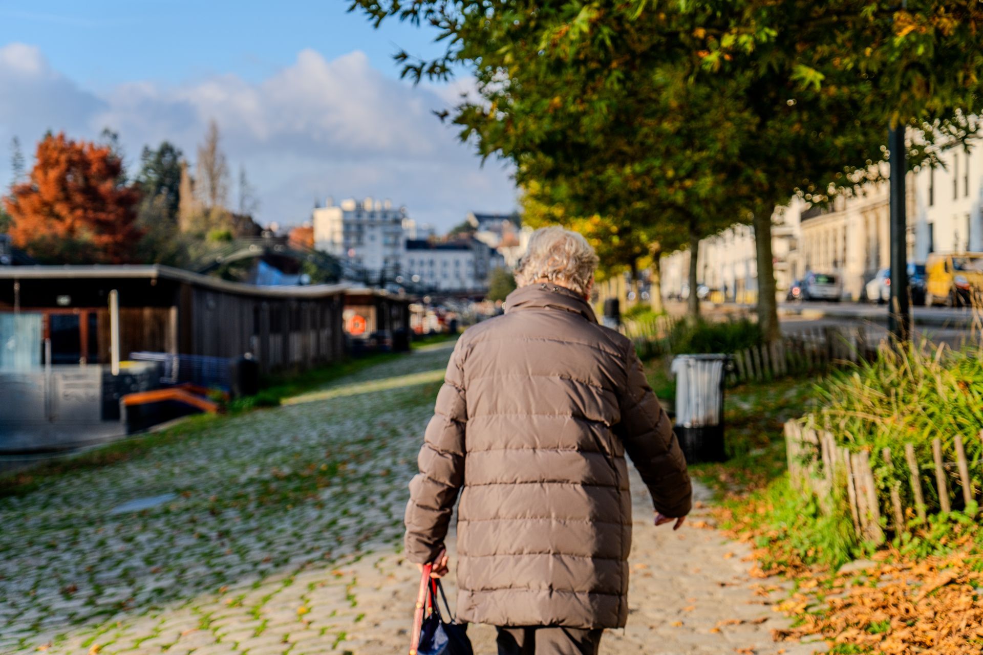 Elderly person in a brown coat walking alone on a cobblestone path in autumn