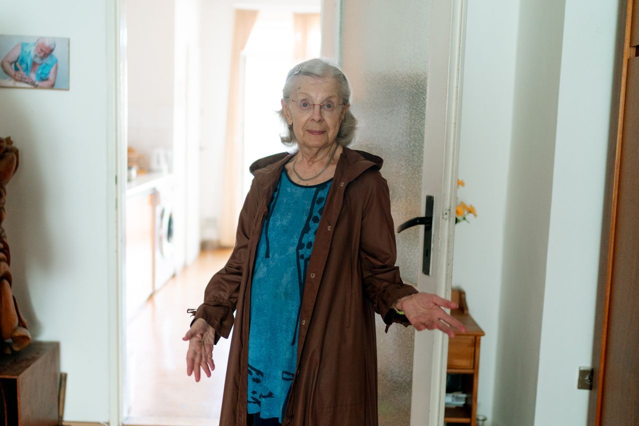 Elderly woman with grey hair standing in a doorway of her home, hands open in a conversational gesture