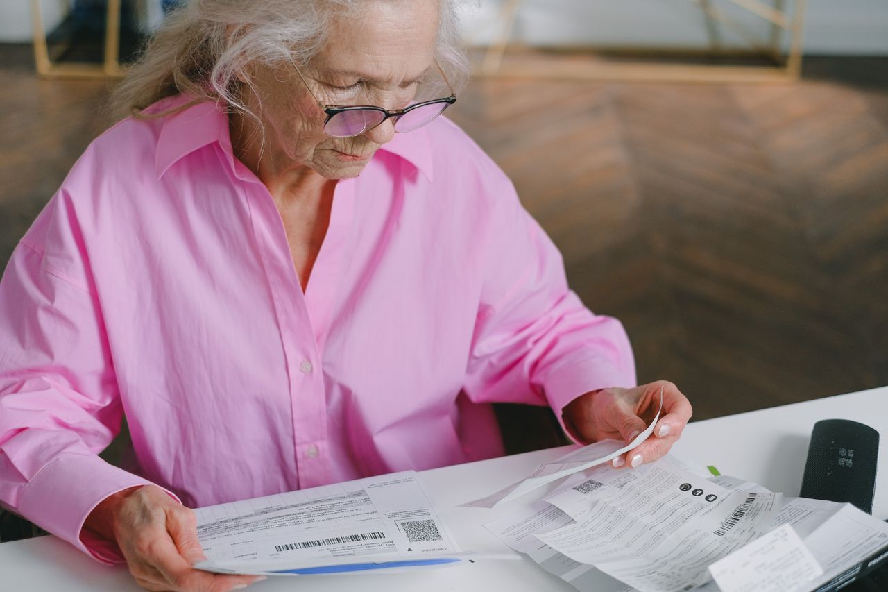 Older woman in glasses reading financial documents at a table indoors
