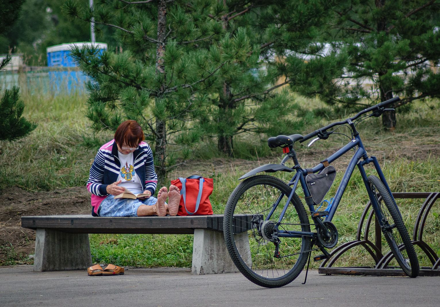 Older woman reading barefoot on a park bench beside her bicycle