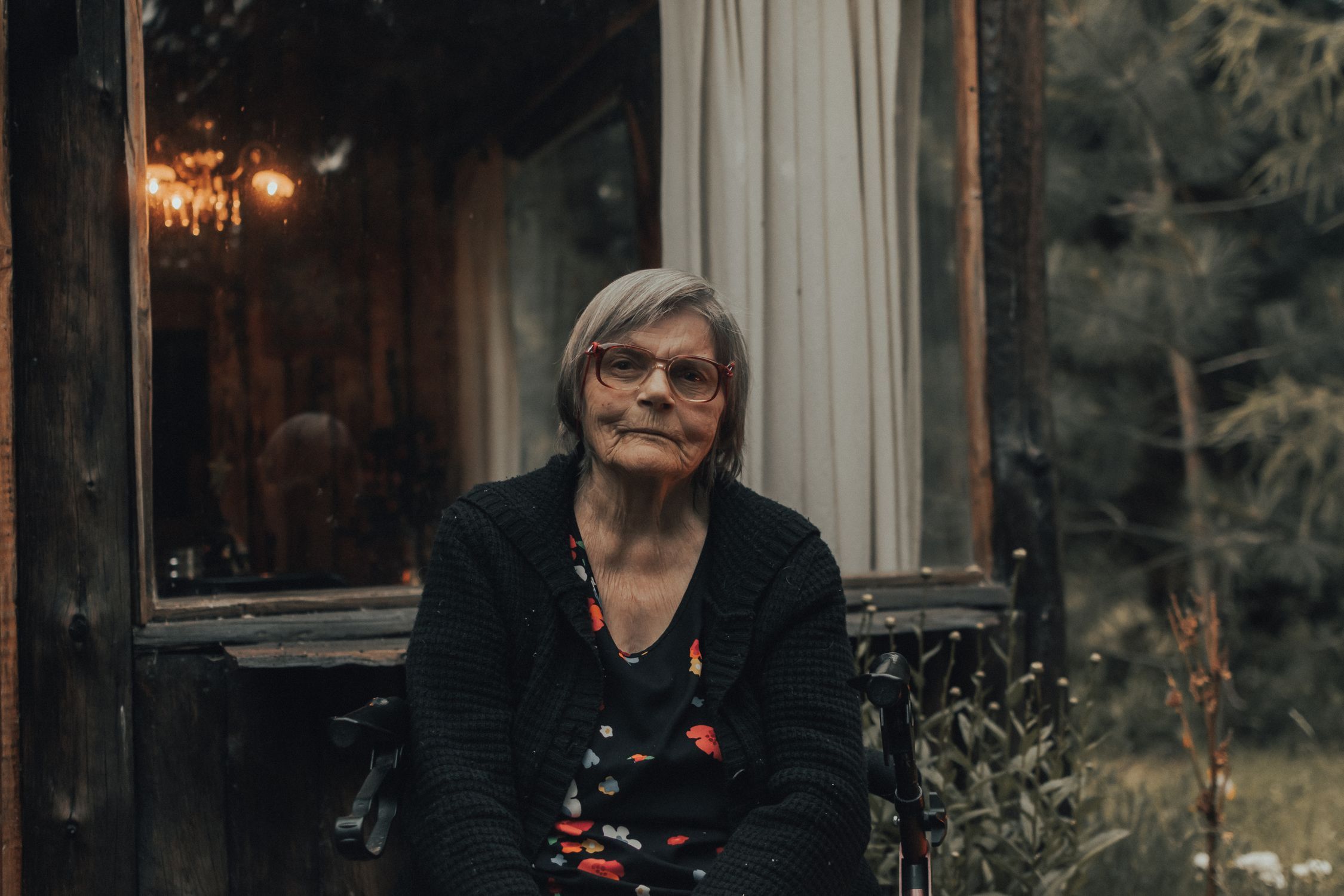 Senior woman with grey hair and glasses sitting outside a rustic cabin surrounded by greenery