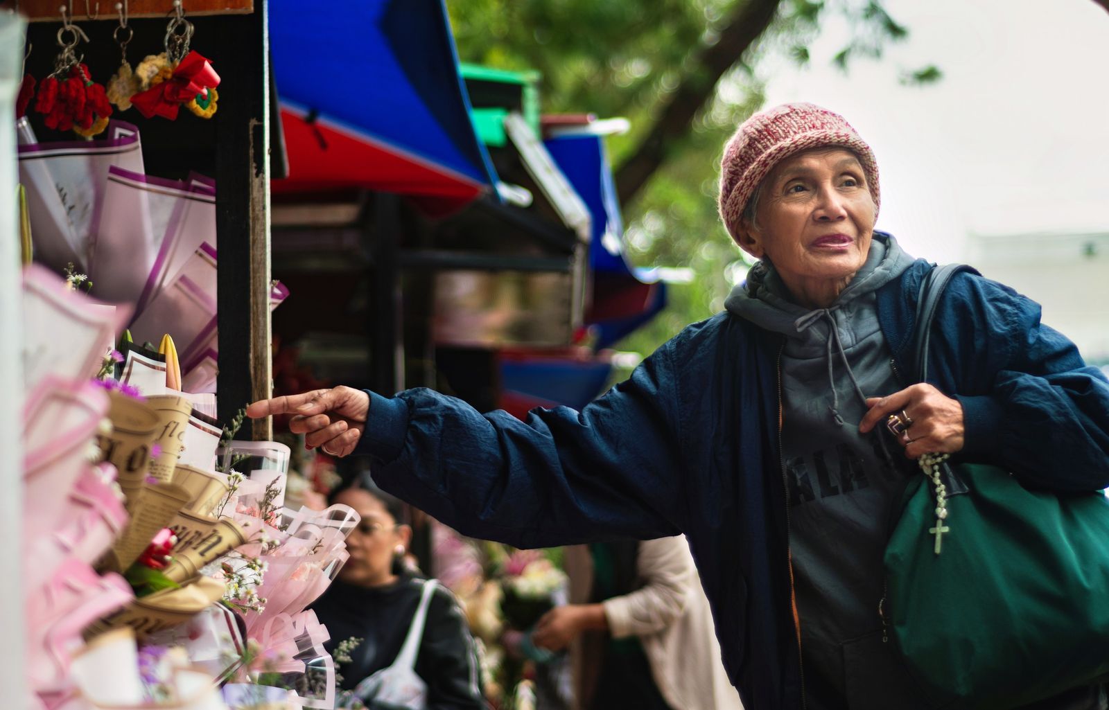 Senior woman in a knit beanie browsing flower bouquets alone at a busy outdoor market stall