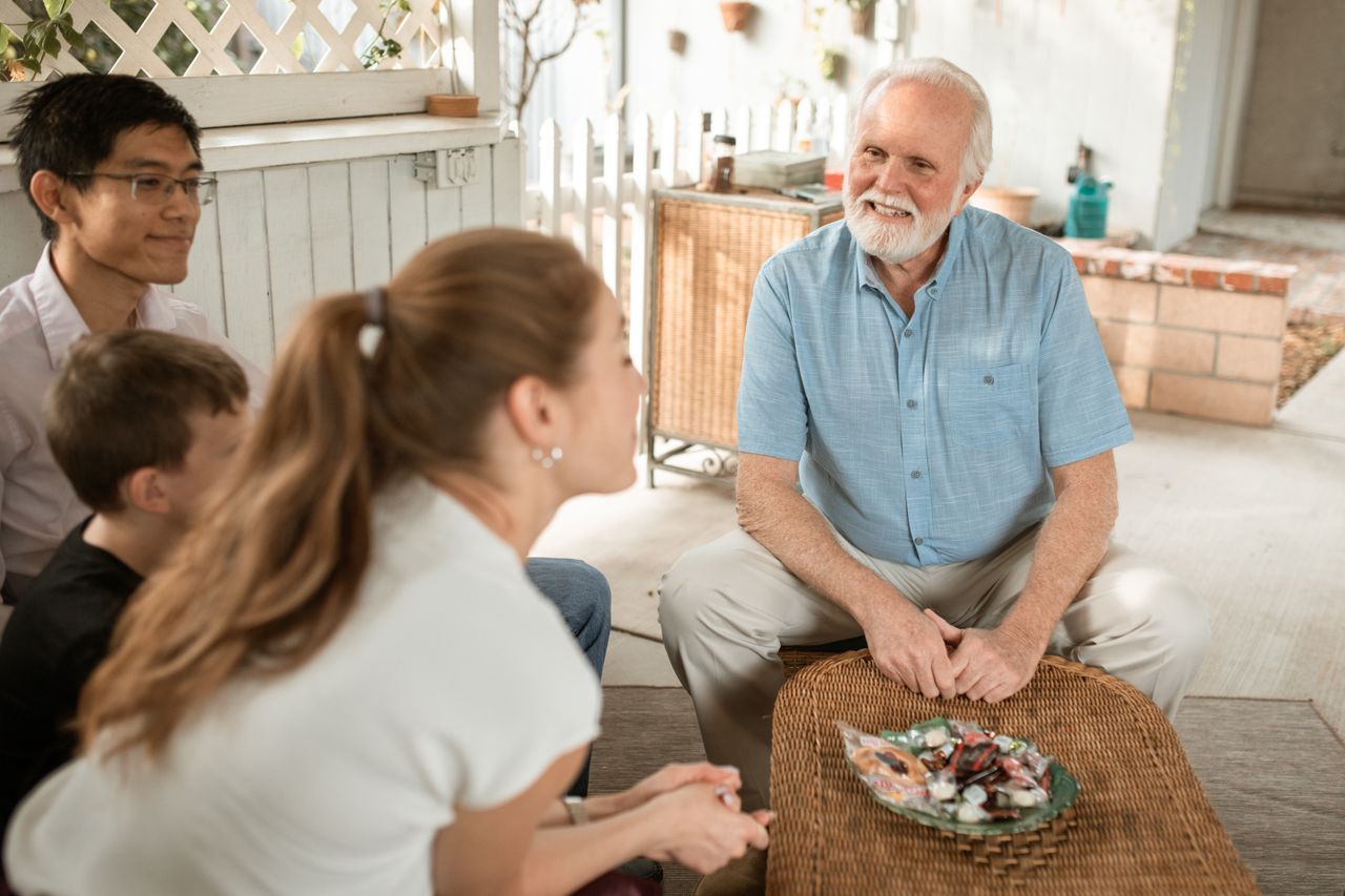 White-bearded older man smiling in conversation with others on a sunny porch
