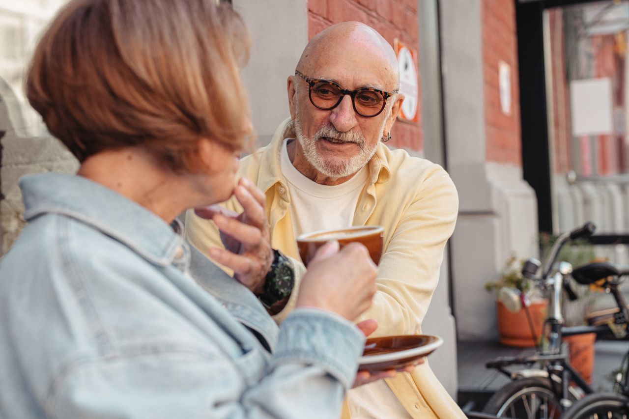 Older man with glasses leaning forward attentively over coffee in conversation with a woman at an outdoor cafe