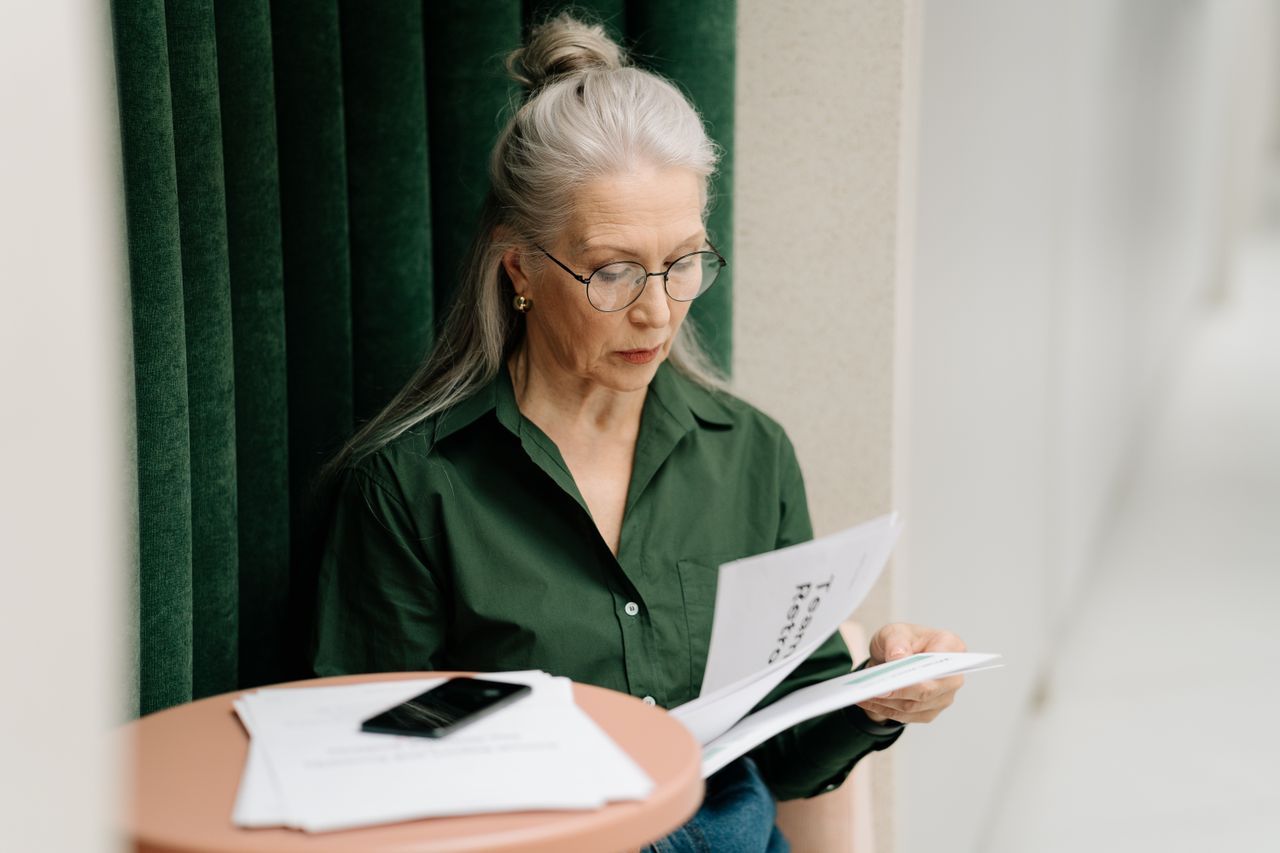 Older woman with grey hair reading papers at a table indoors in soft light