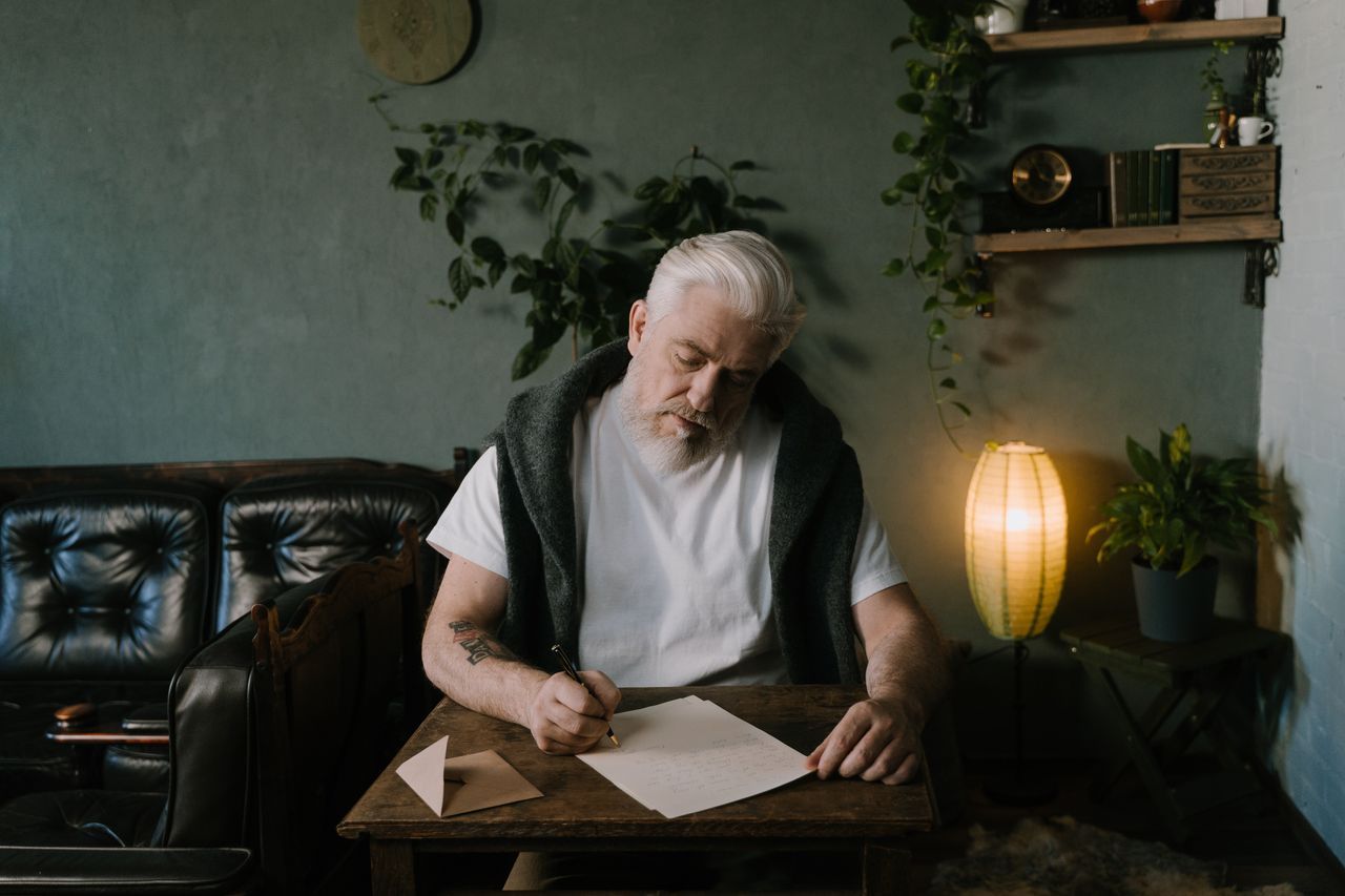 Senior man with white hair writing at a wooden table in warm home light