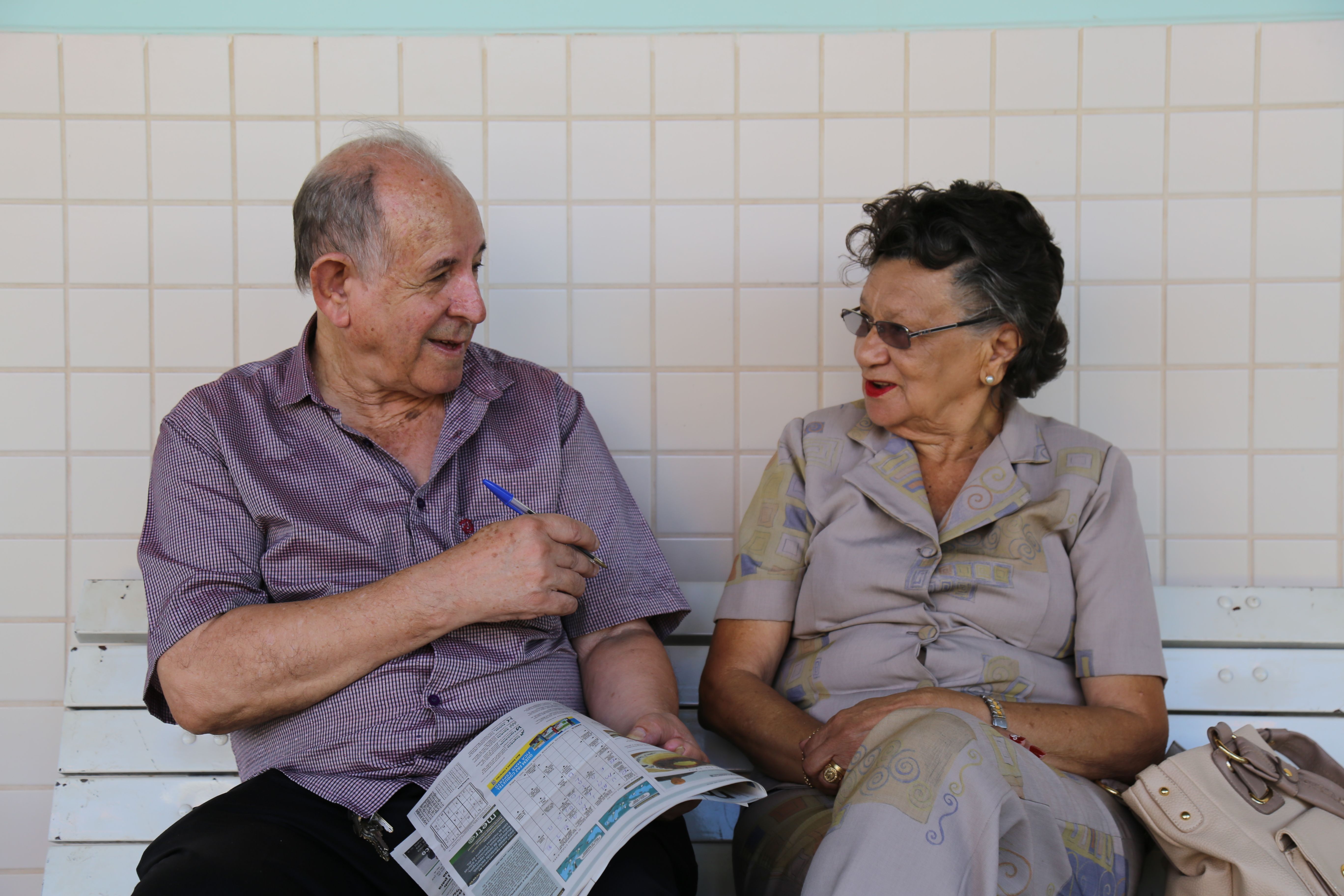 Older man and woman in conversation on a white bench with a newspaper between them