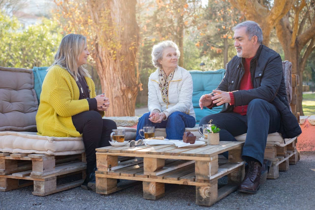 Three older women chatting over tea at a sunny park table surrounded by trees