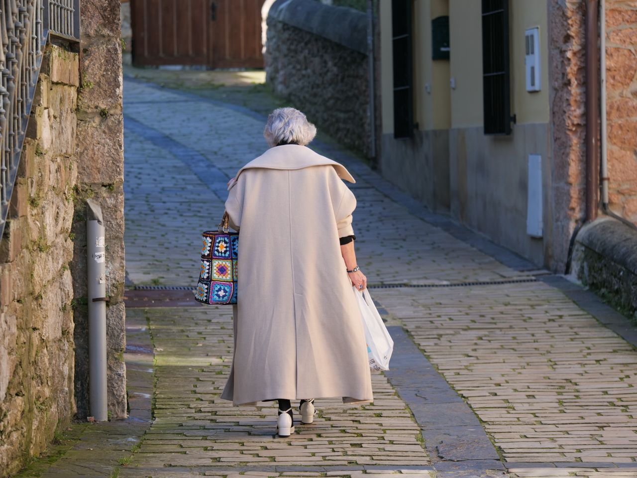 Grey-haired woman in a beige overcoat walking alone down a quiet cobblestone alley in warm afternoon light