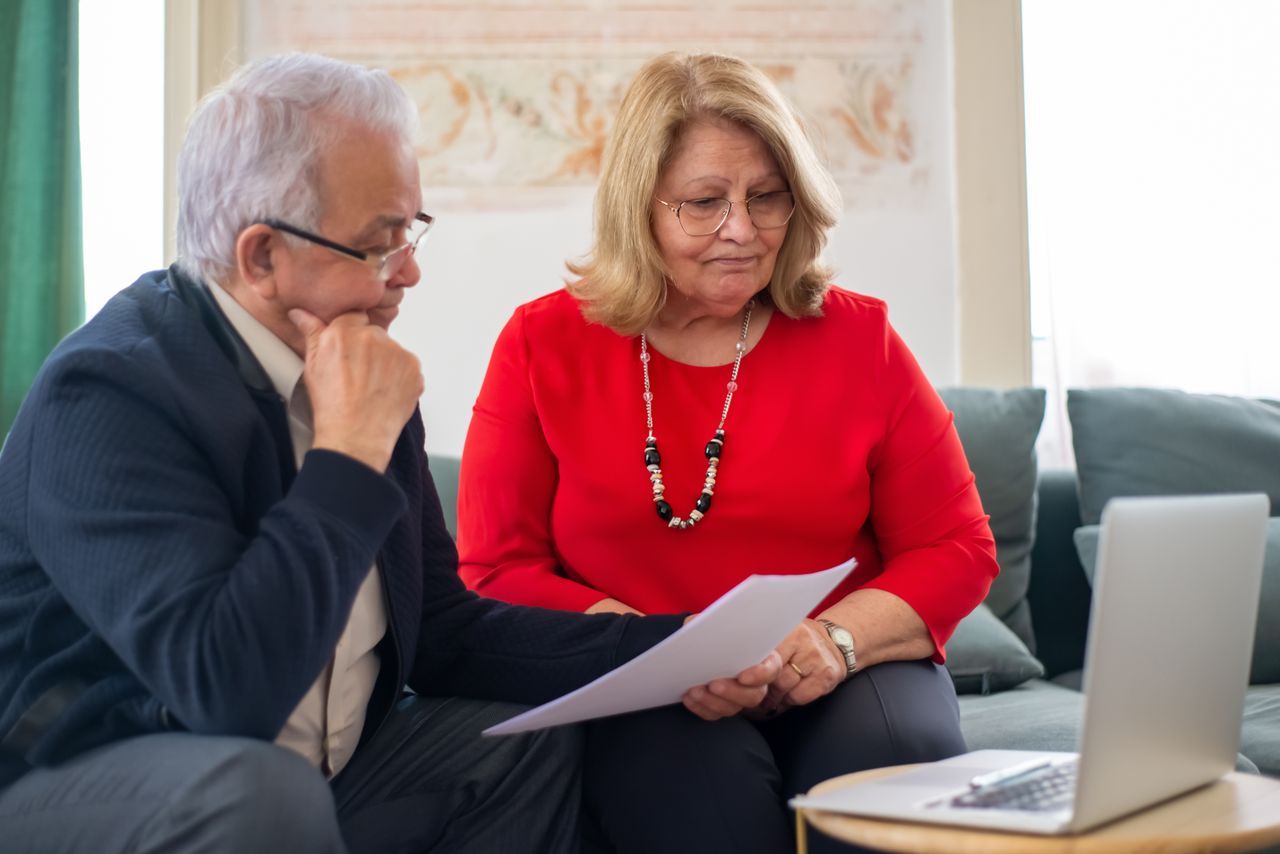 Older couple with grey hair reviewing a document together on a sofa in a warm living room