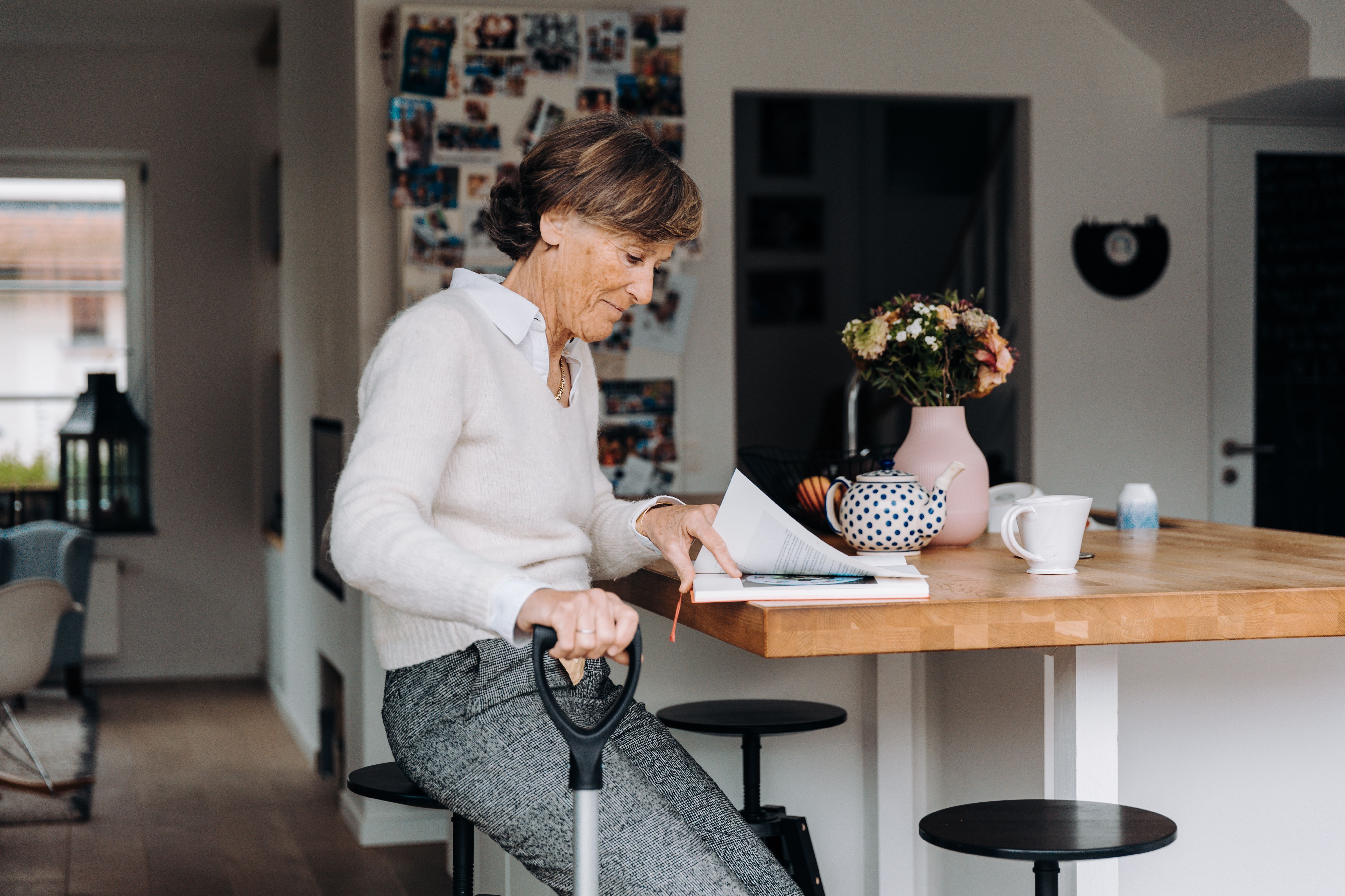 Senior woman with grey hair reading at a kitchen table in a cozy home with flowers and warm light