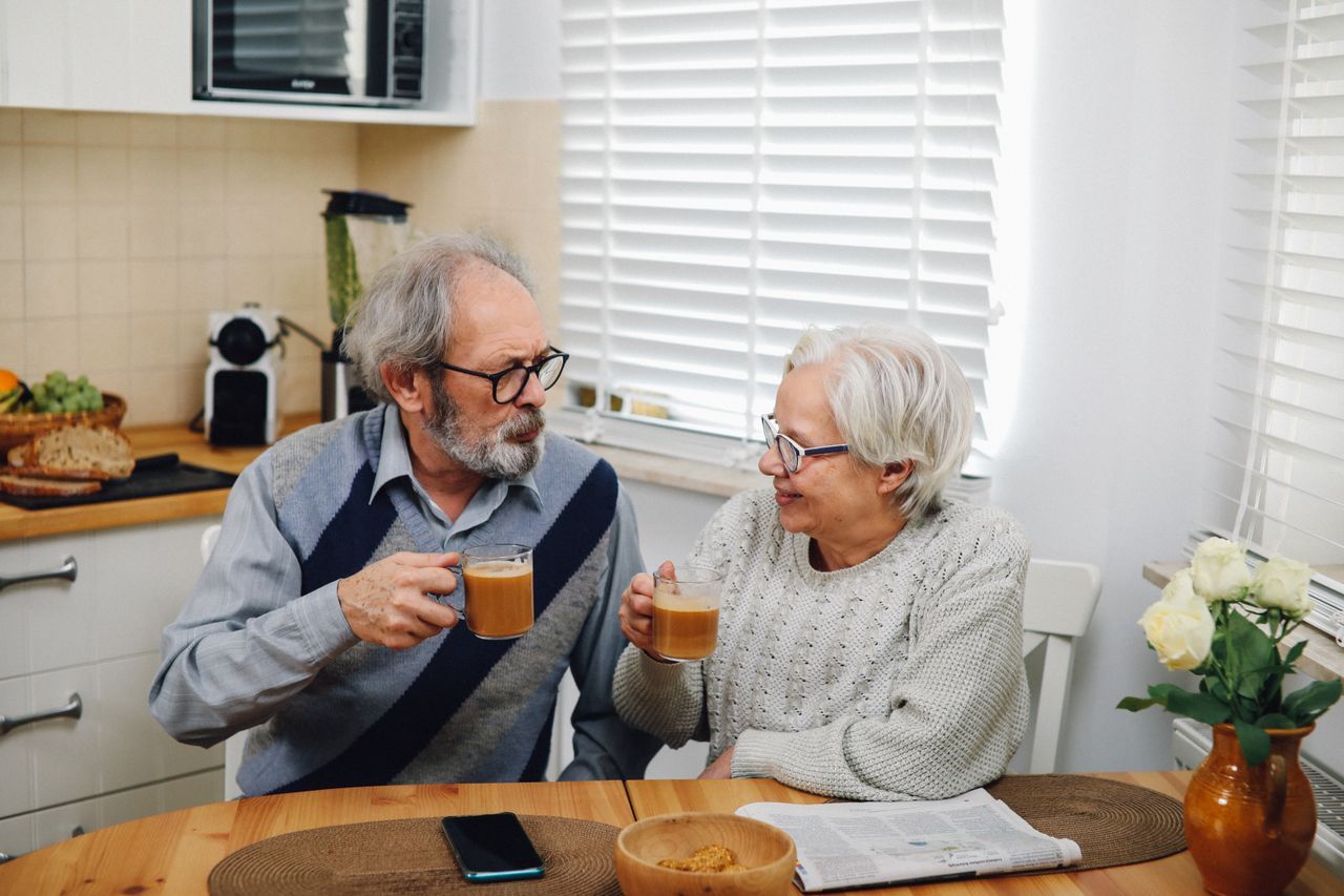Older couple with grey hair sharing coffee at a kitchen table, looking at each other in quiet conversation