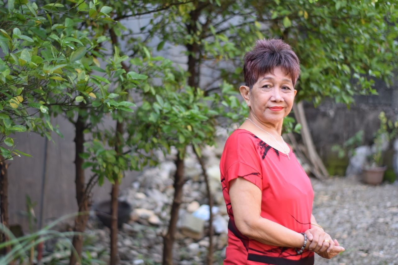 Mature woman standing alone in a garden with green foliage, wearing a red top and looking quietly content