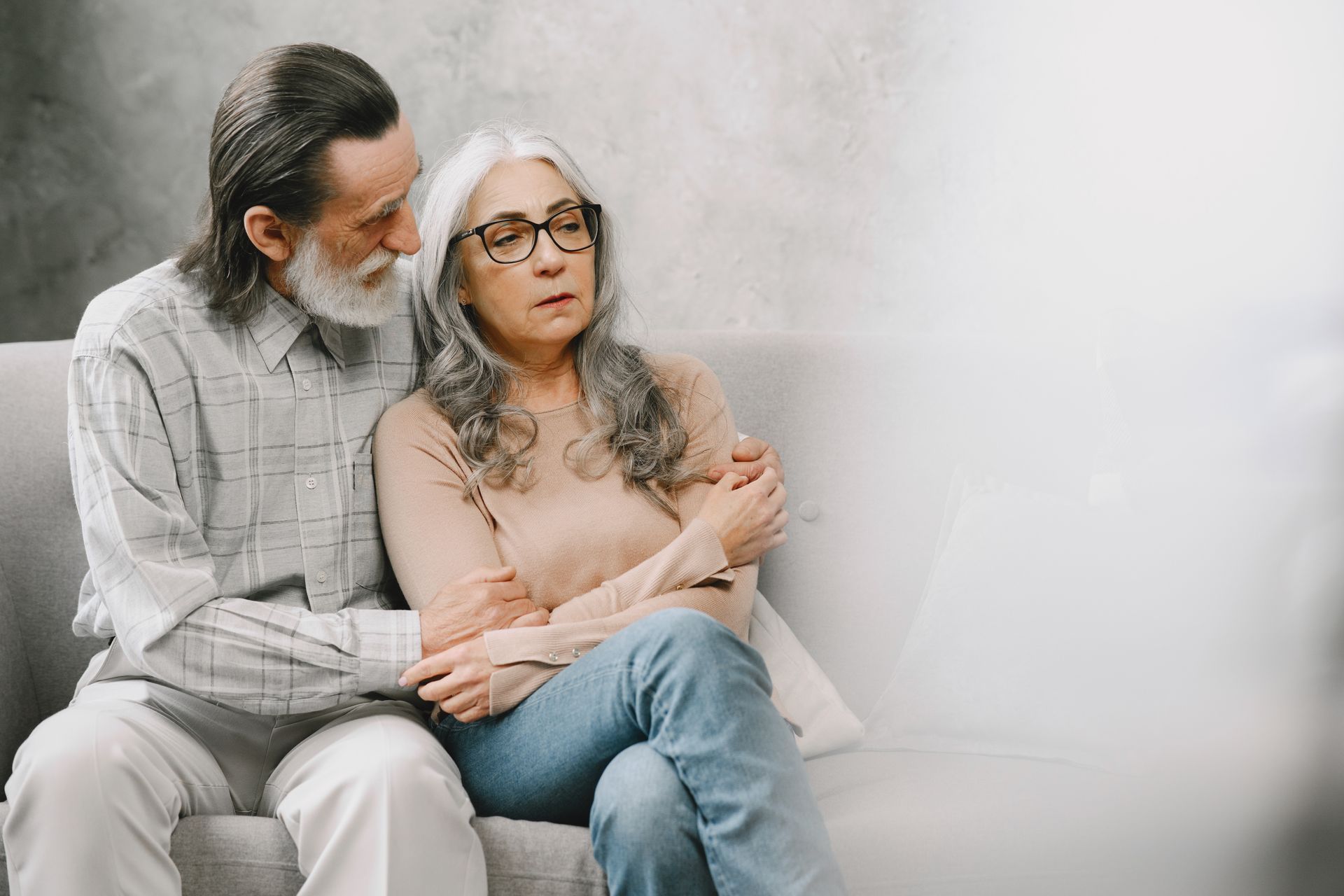 Senior couple sitting close on a sofa, one comforting the other in warm indoor light