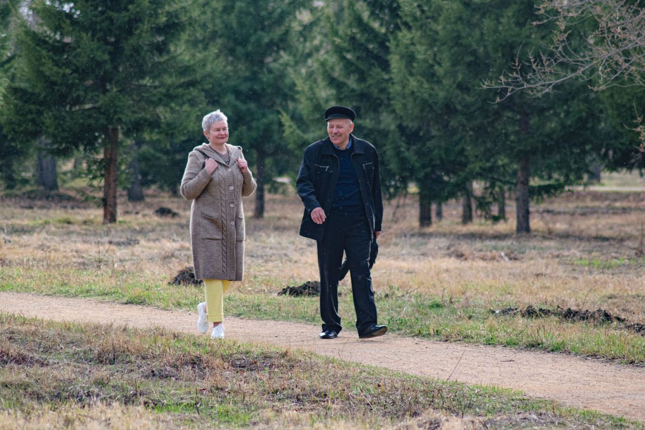 Older couple walking together on a dirt path through a park with evergreen trees