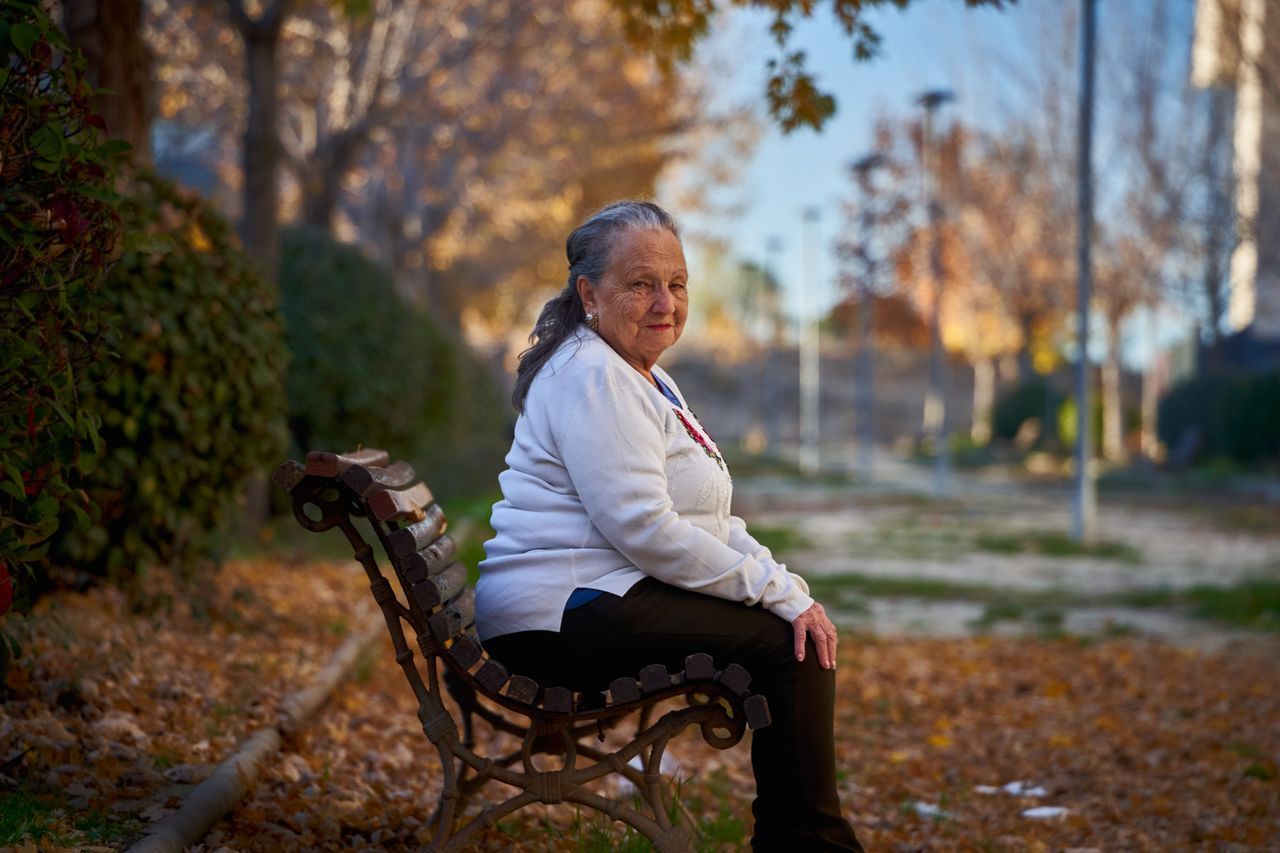 Grey-haired woman in her seventies sitting on an iron bench in an autumn park with fallen leaves