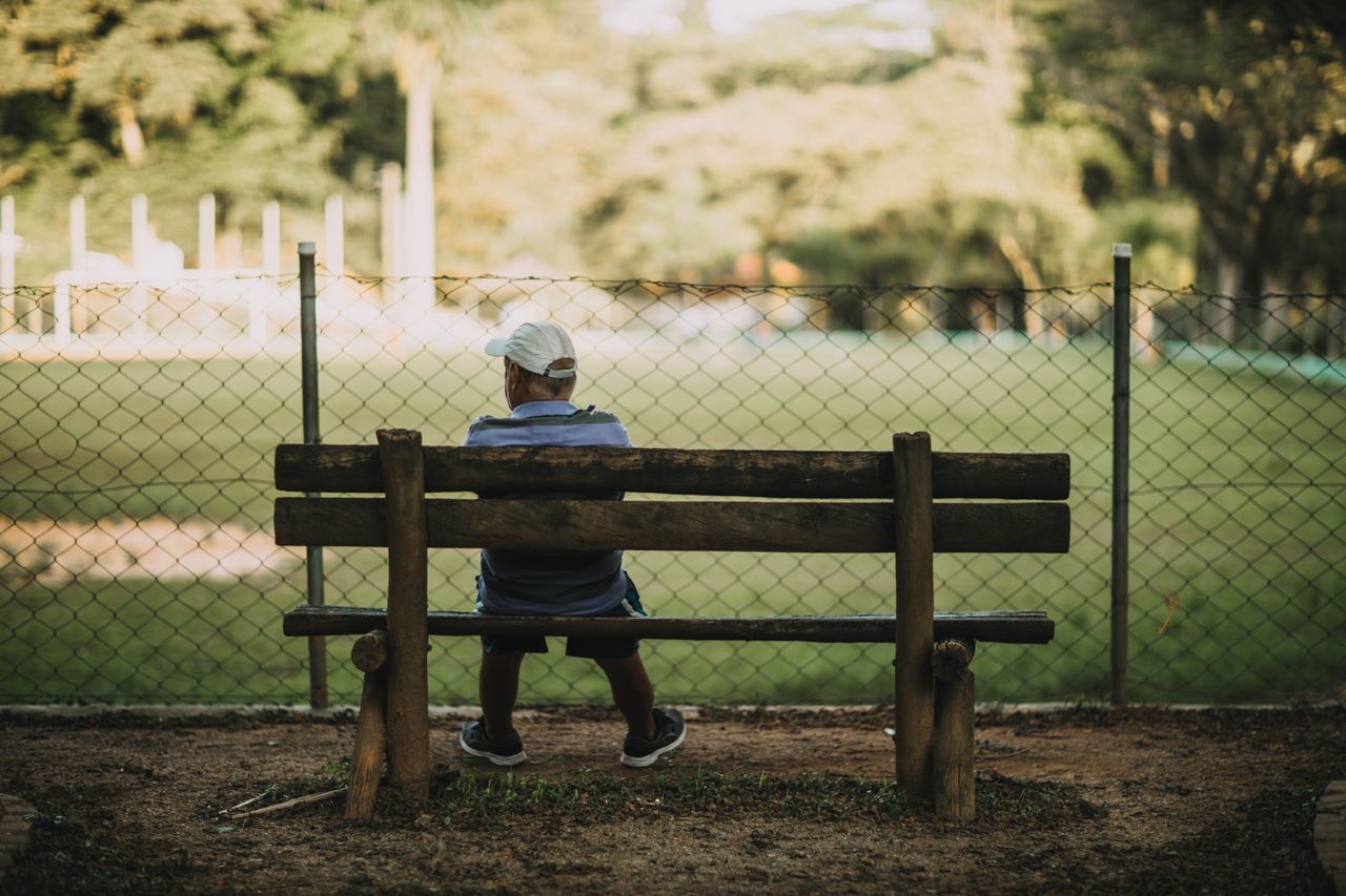 Senior man in white cap sitting alone on a wooden park bench, seen from behind