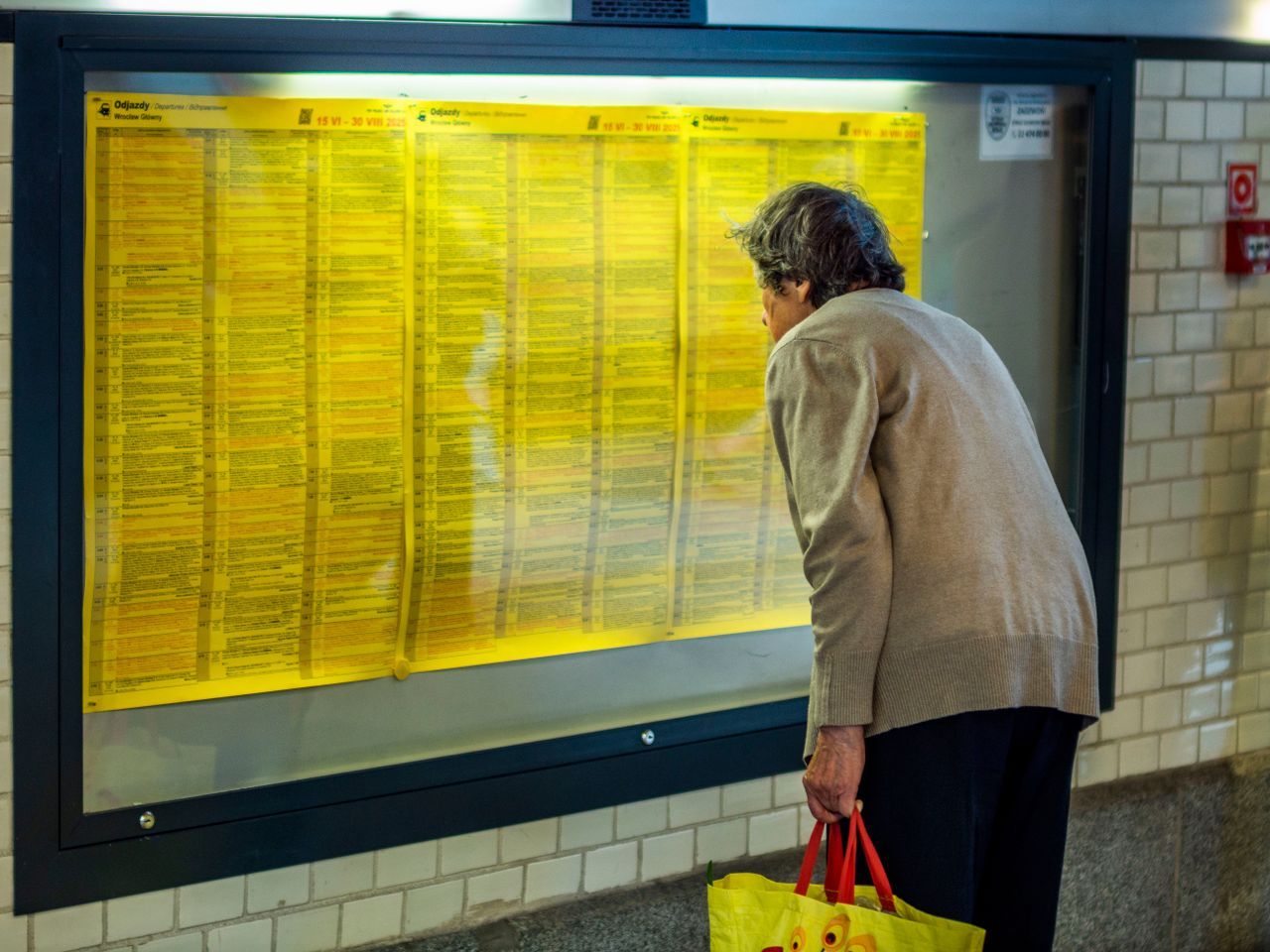 Older woman reading a train departure timetable at a station, carrying a shopping bag