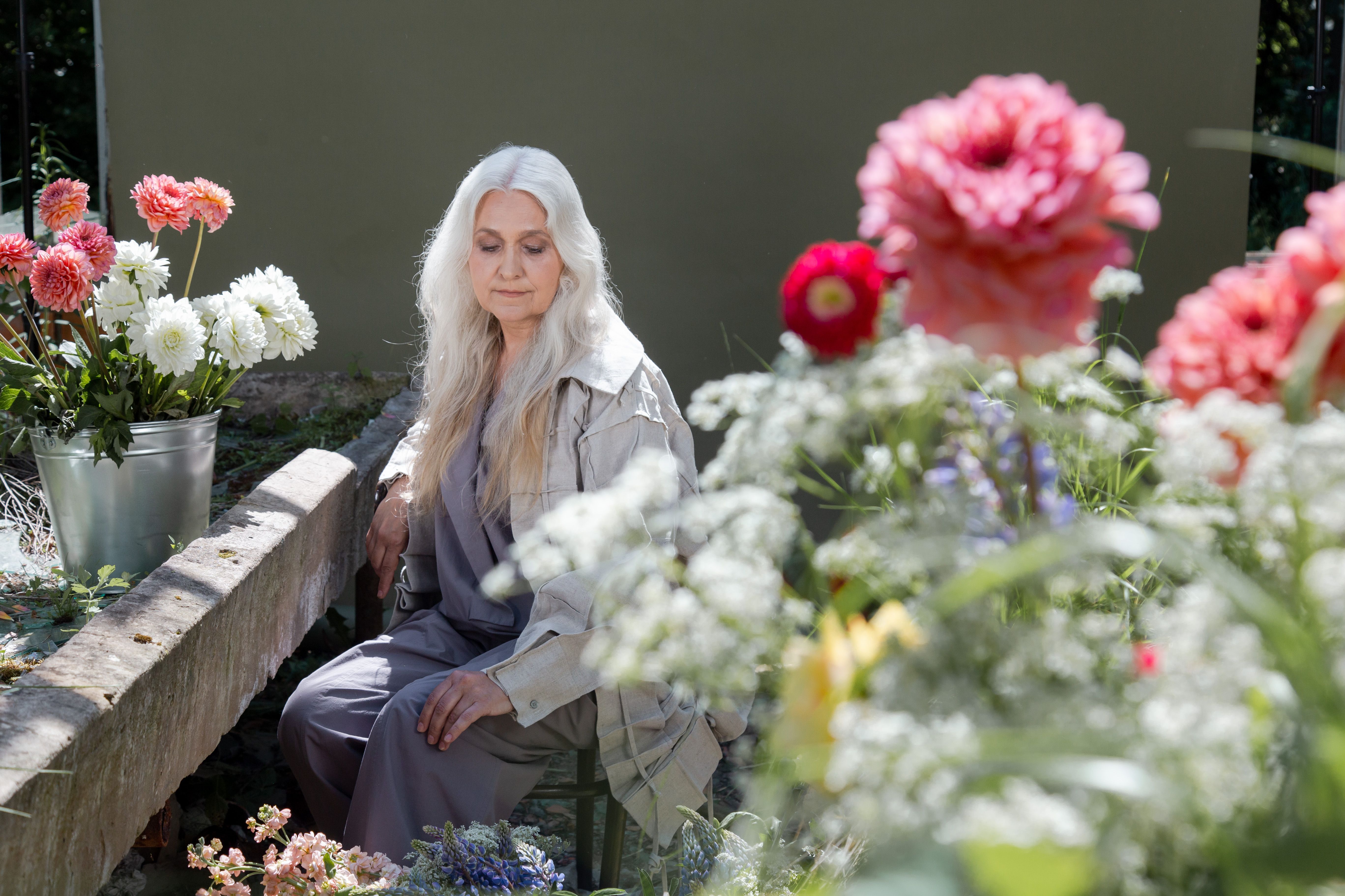 Grey-haired woman sitting thoughtfully among flowers in a garden