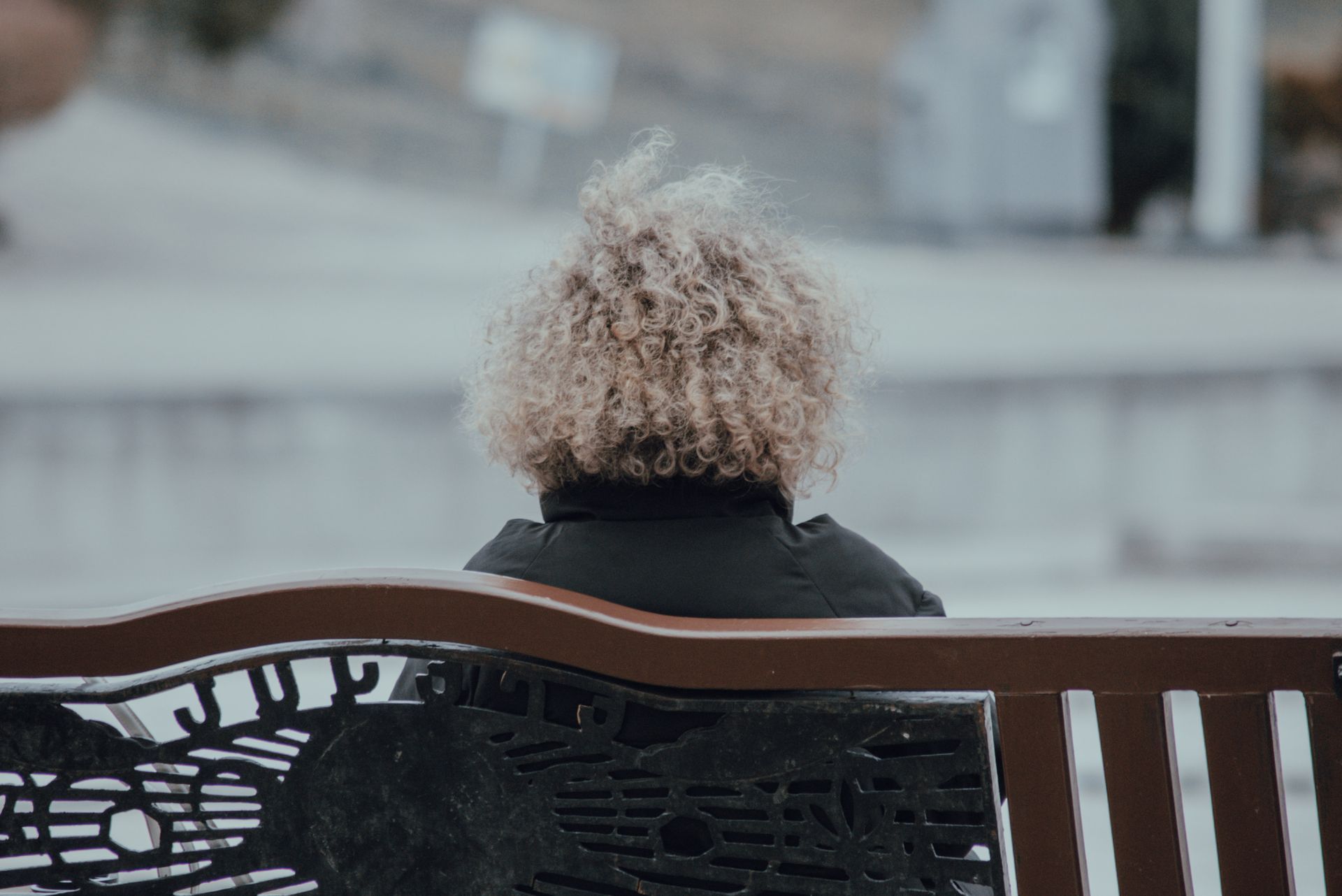 Grey-haired woman sitting alone on a bench in a city, seen from behind