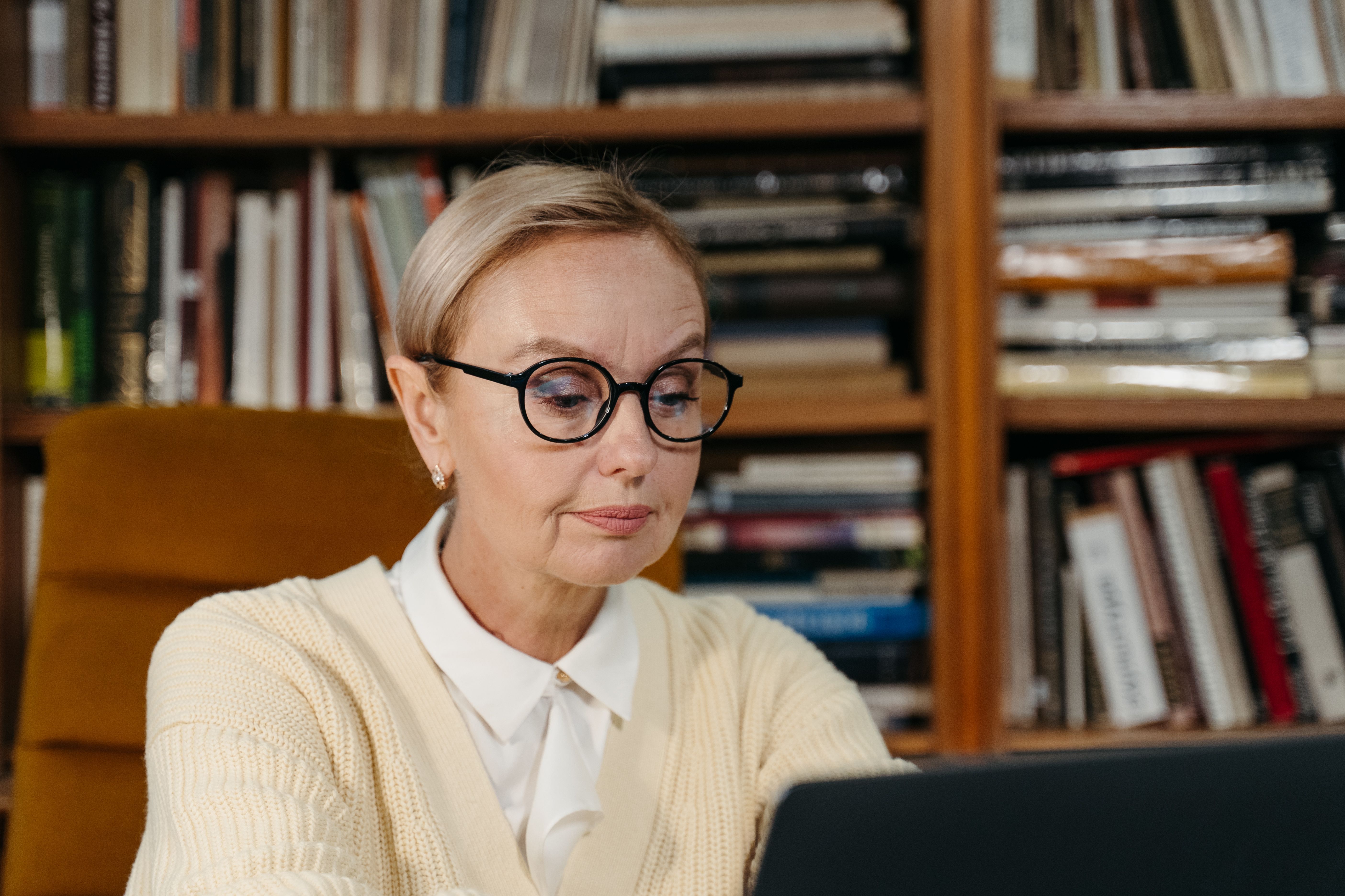Woman in her 60s with glasses looking at laptop in home library with bookshelves