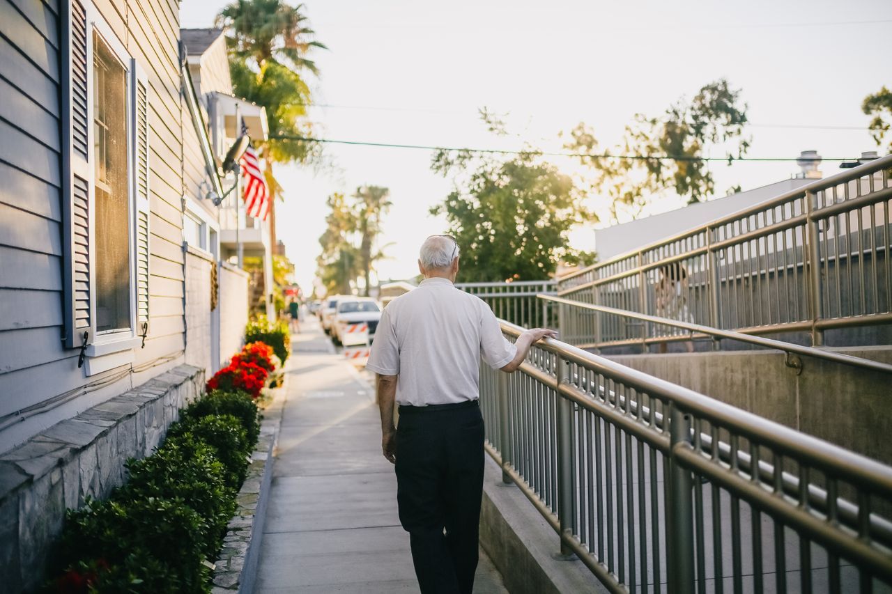 Elderly man with white hair walking along a sunny residential pathway past houses and flowers