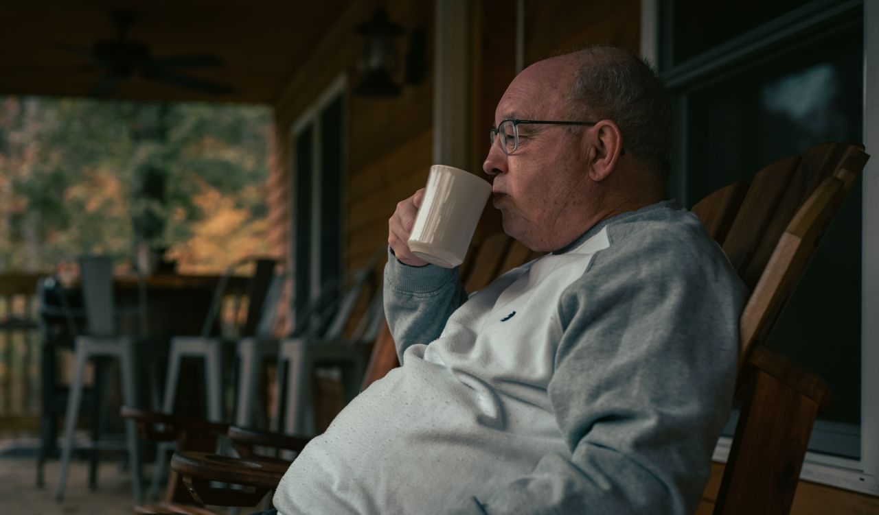Senior man in glasses drinking from a mug on a covered porch in autumn light