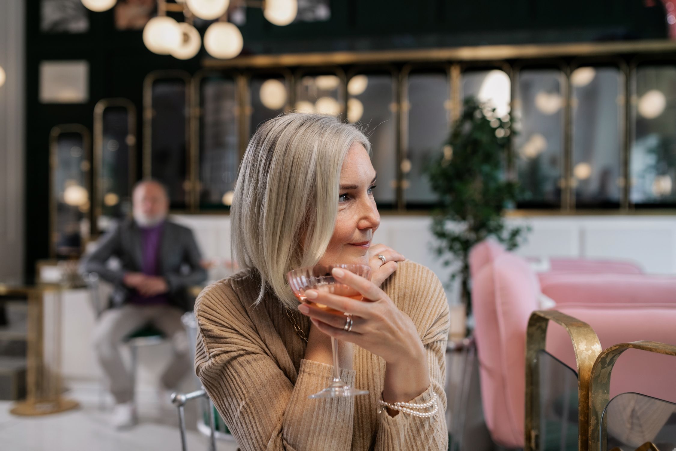 Woman in her early 60s holding a wine glass in a stylish cafe, looking contemplative