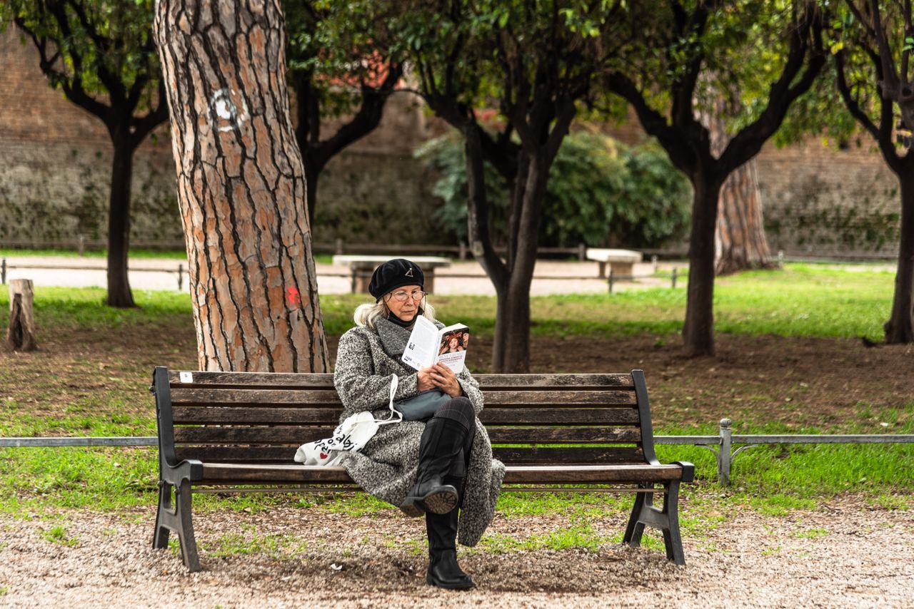 Older woman reading alone on a park bench surrounded by trees and greenery